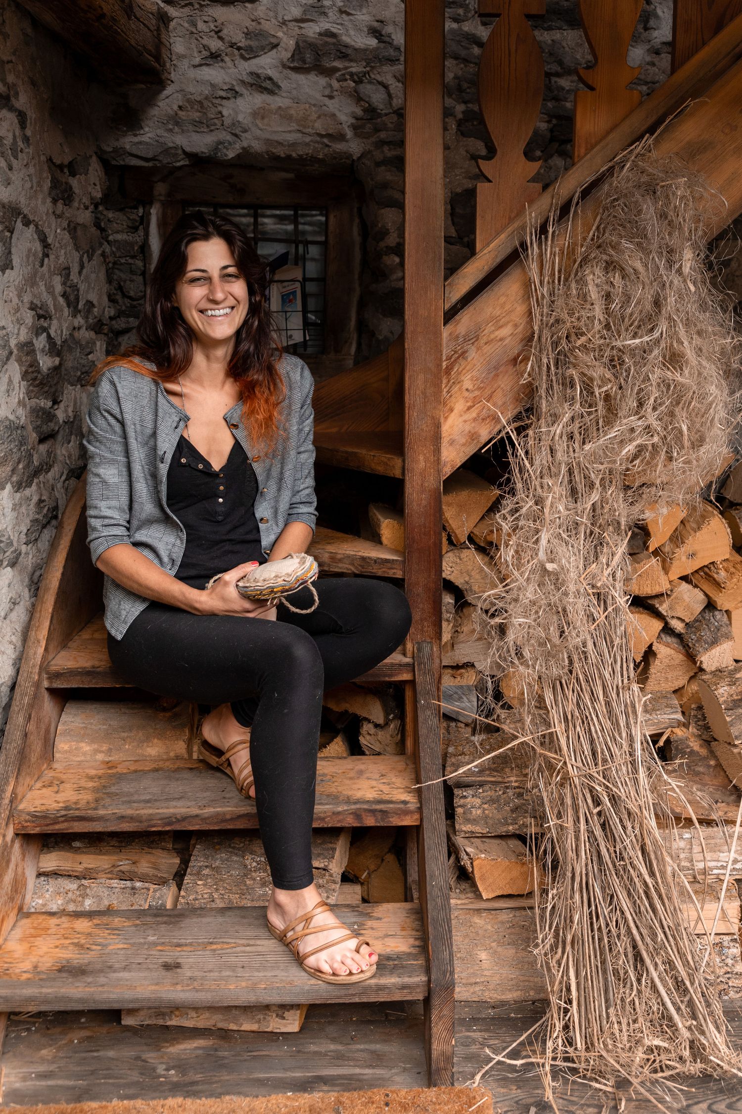 A smiling woman sits on wooden stairs beside stacked firewood in a stone building.