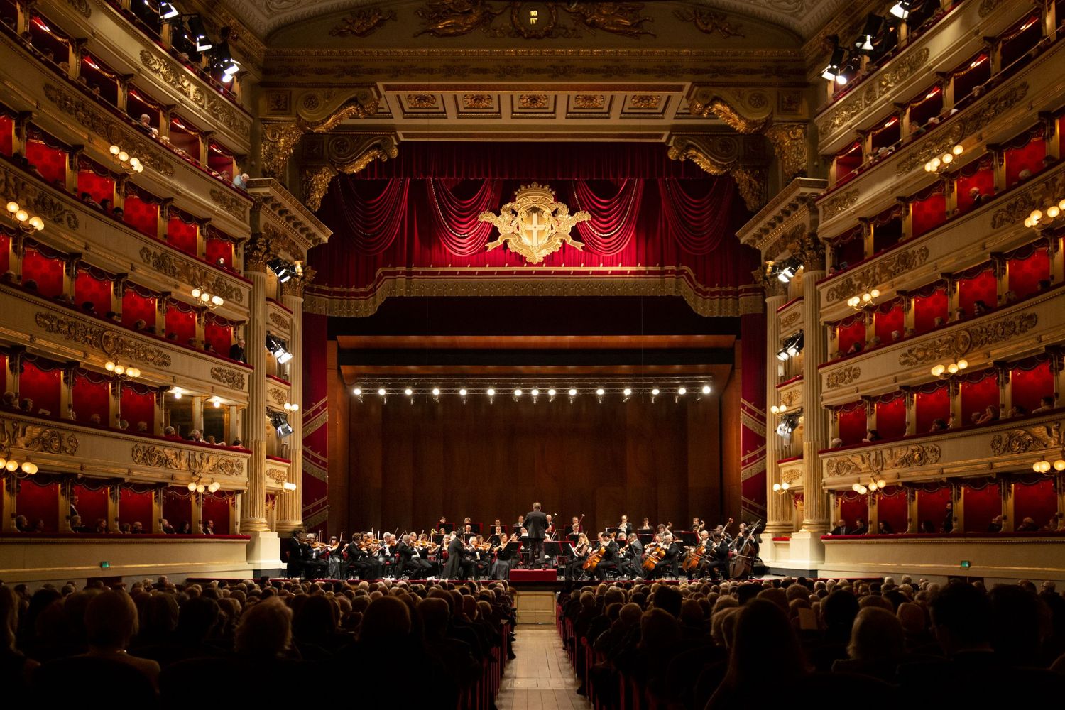 Grand interior of La Scala opera house with orchestra on stage and ornate red tiered balconies.