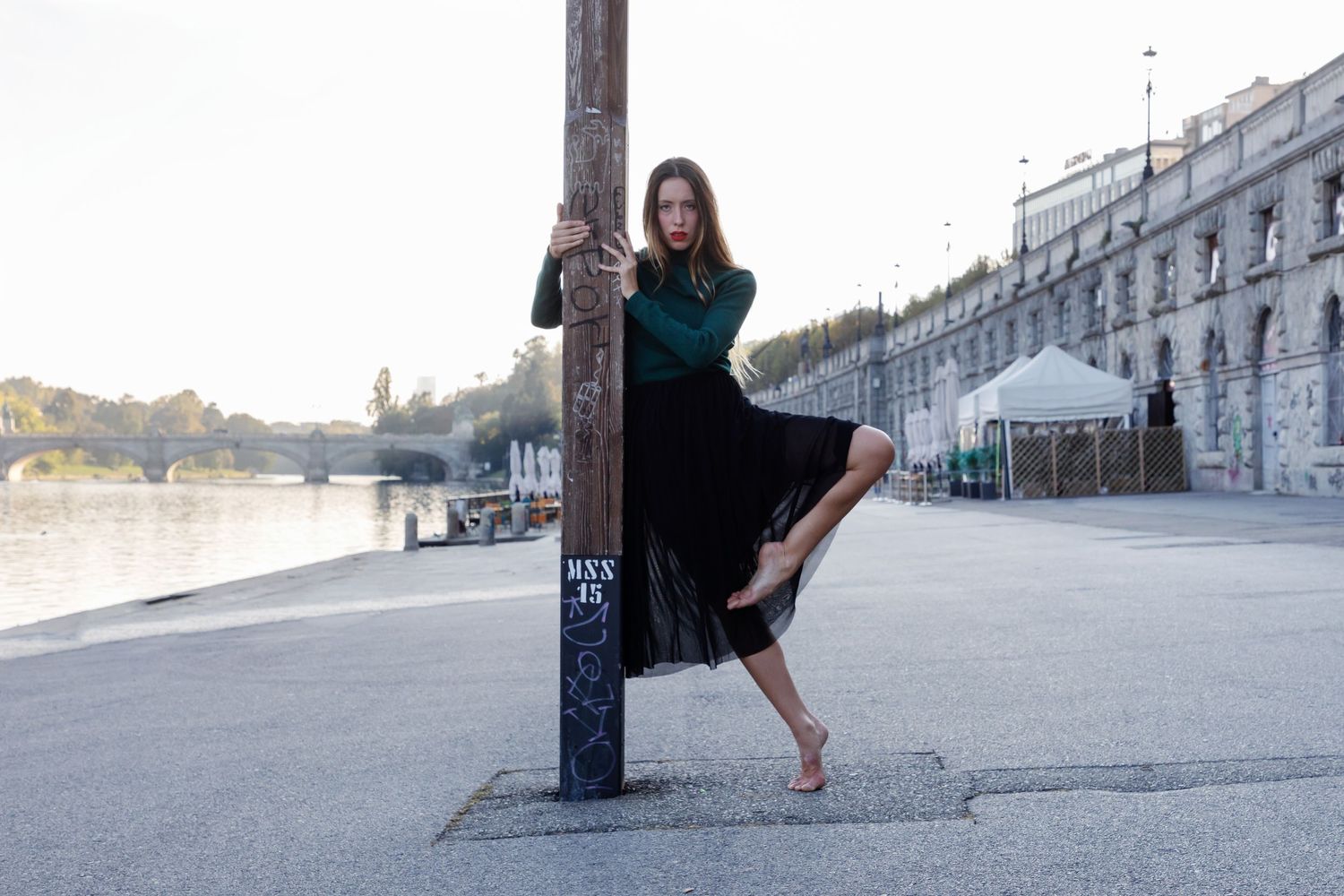 Young woman in black skirt and green top holding wooden pole beside urban riverside promenade.