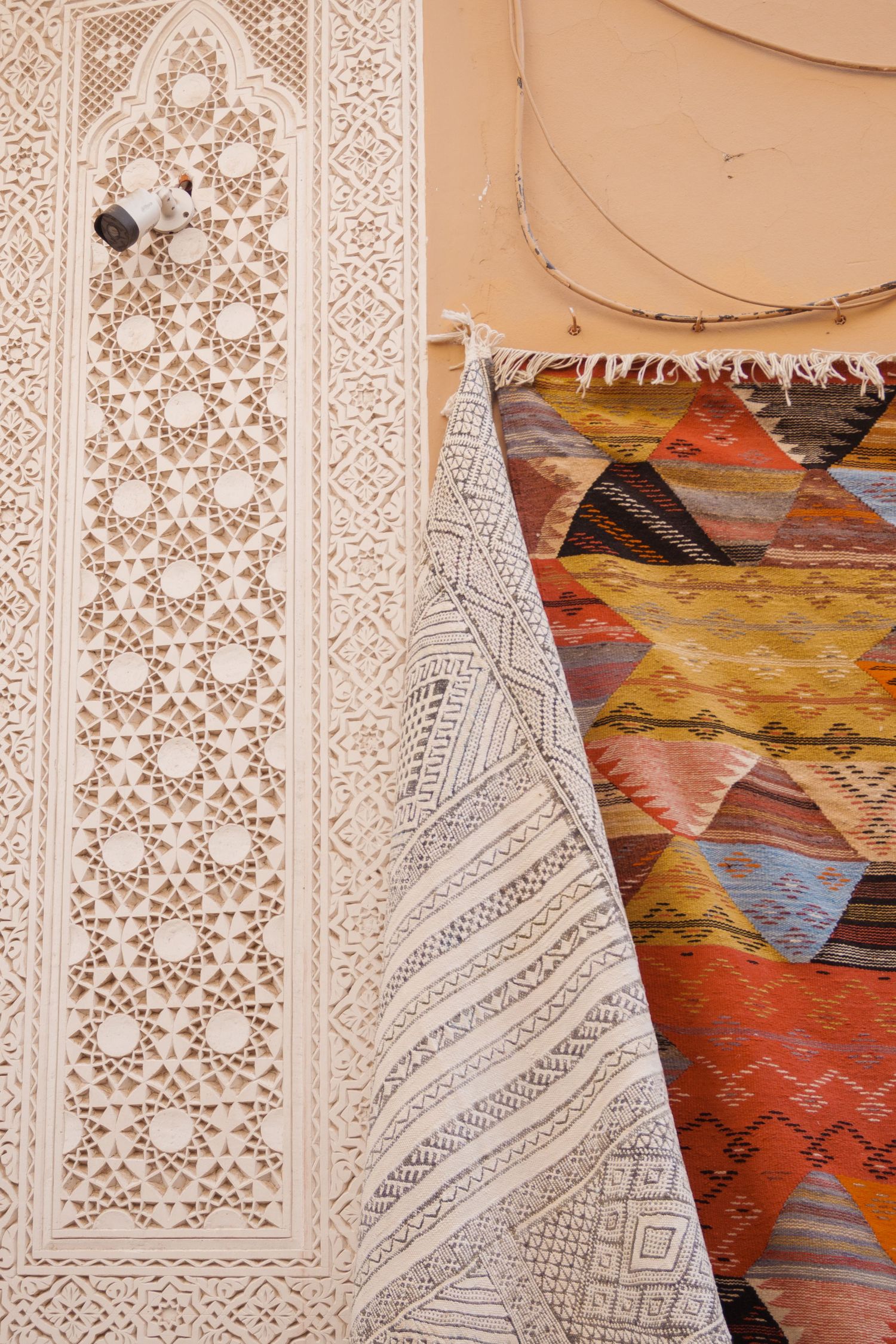 Intricate white Moroccan plasterwork beside colorful geometric patterned rugs hanging on a terracotta wall.
