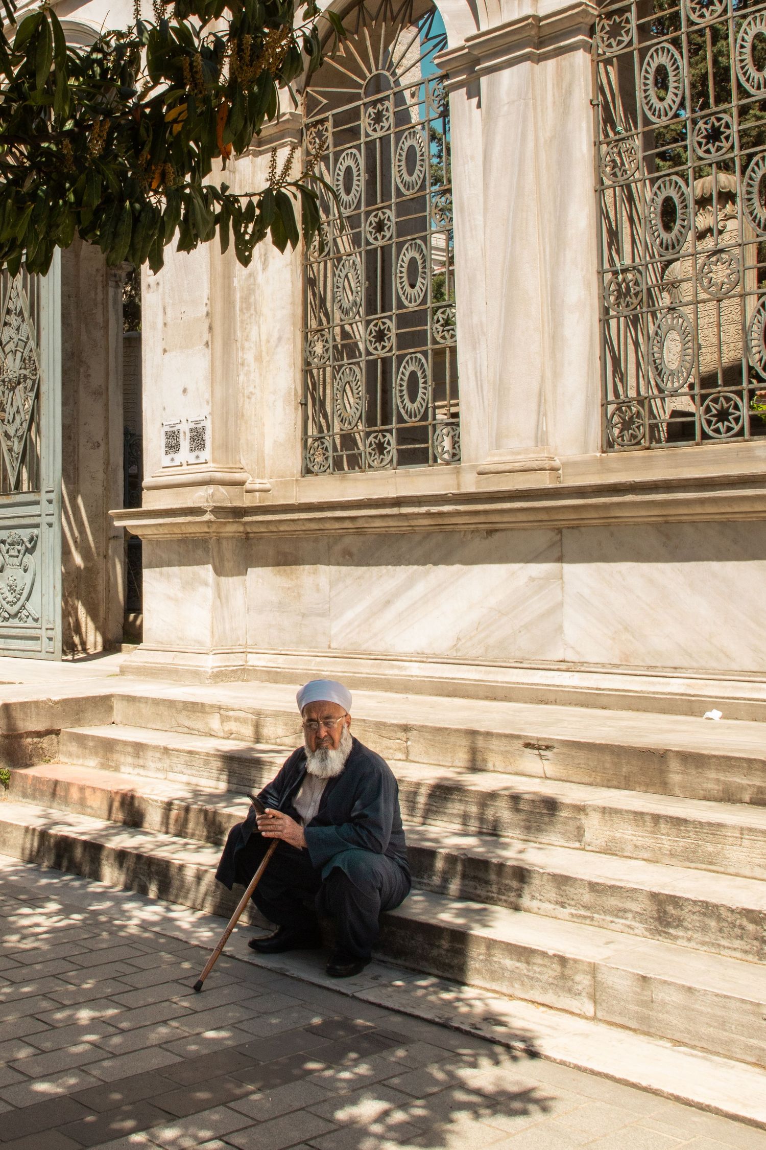 An elderly bearded man in a prayer cap sitting on mosque steps with a walking cane in Istanbul.