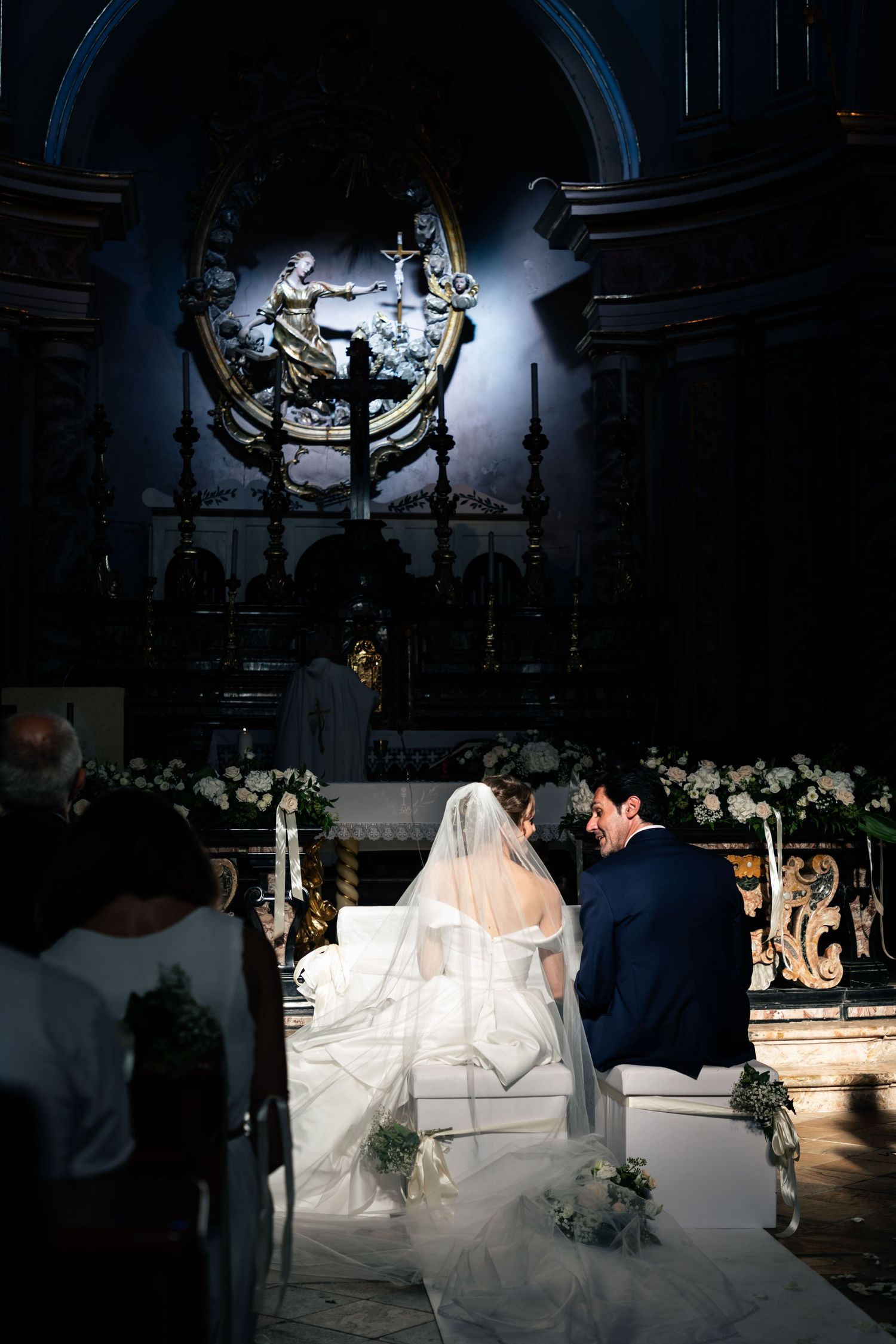 Bride and groom kneel at ornate church altar during wedding ceremony in dimly lit historic cathedral.