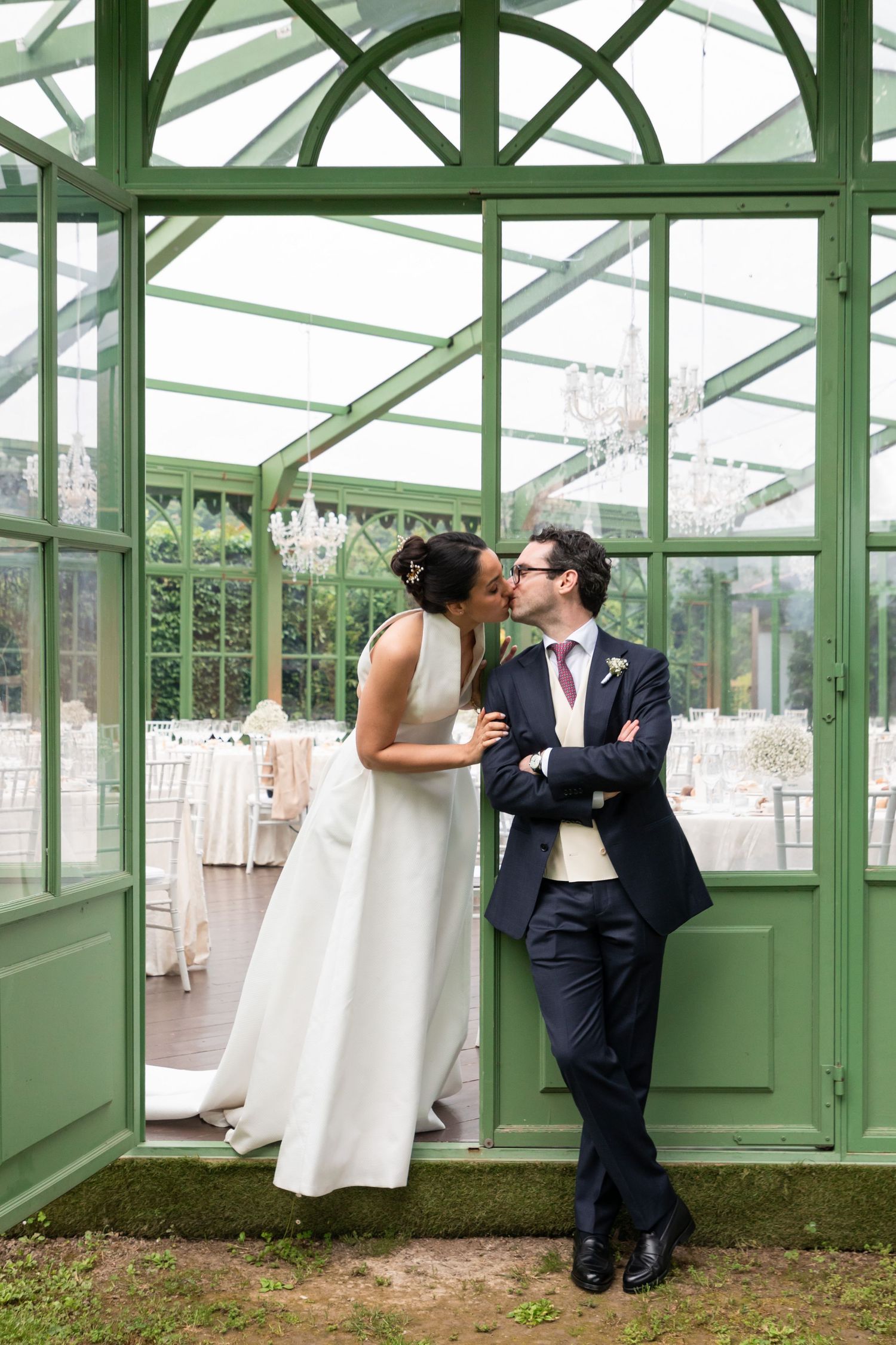 Bride and groom sharing a kiss in doorway of elegant green glass greenhouse wedding venue.
