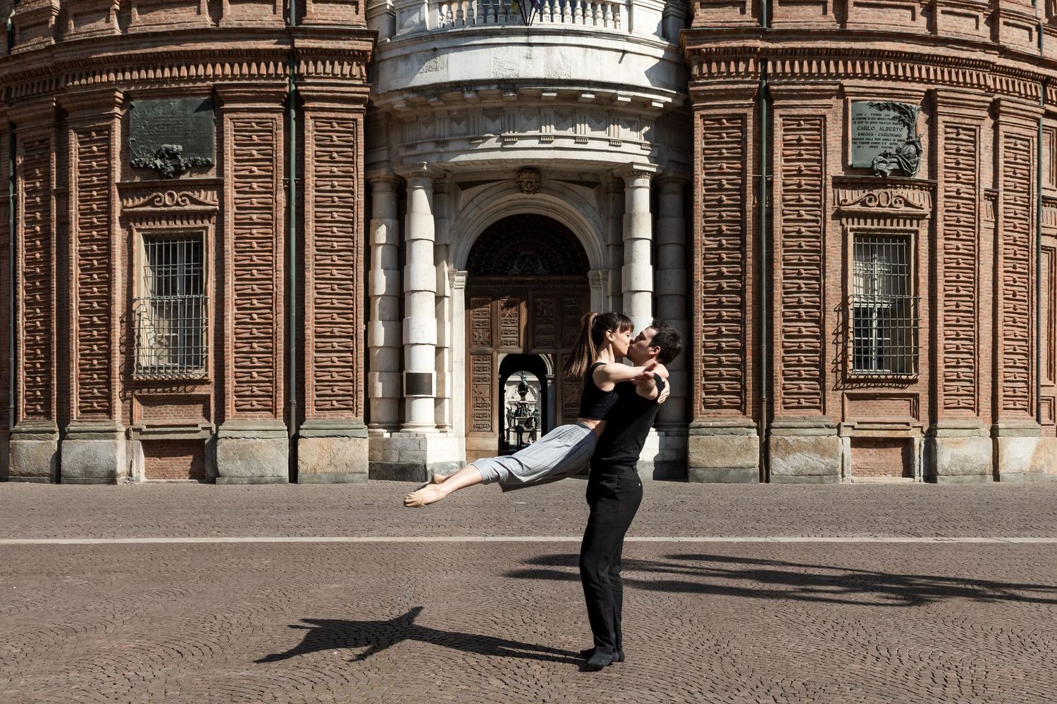 Ballet dancers perform elegant lift outside historic ornate brick building in Turin, Italy.