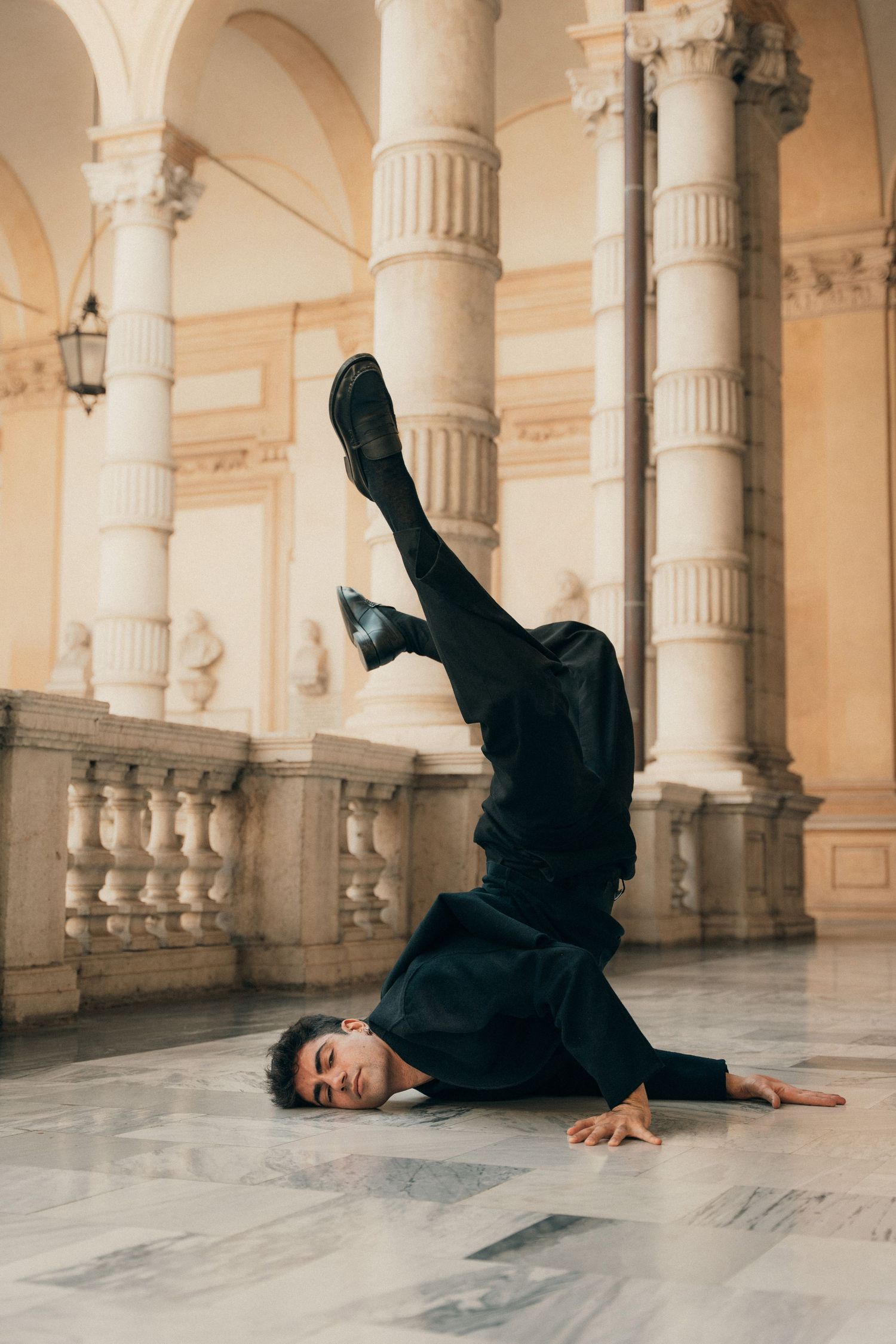 Male dancer in all black performing floor acrobatic move in ornate classical arcade.