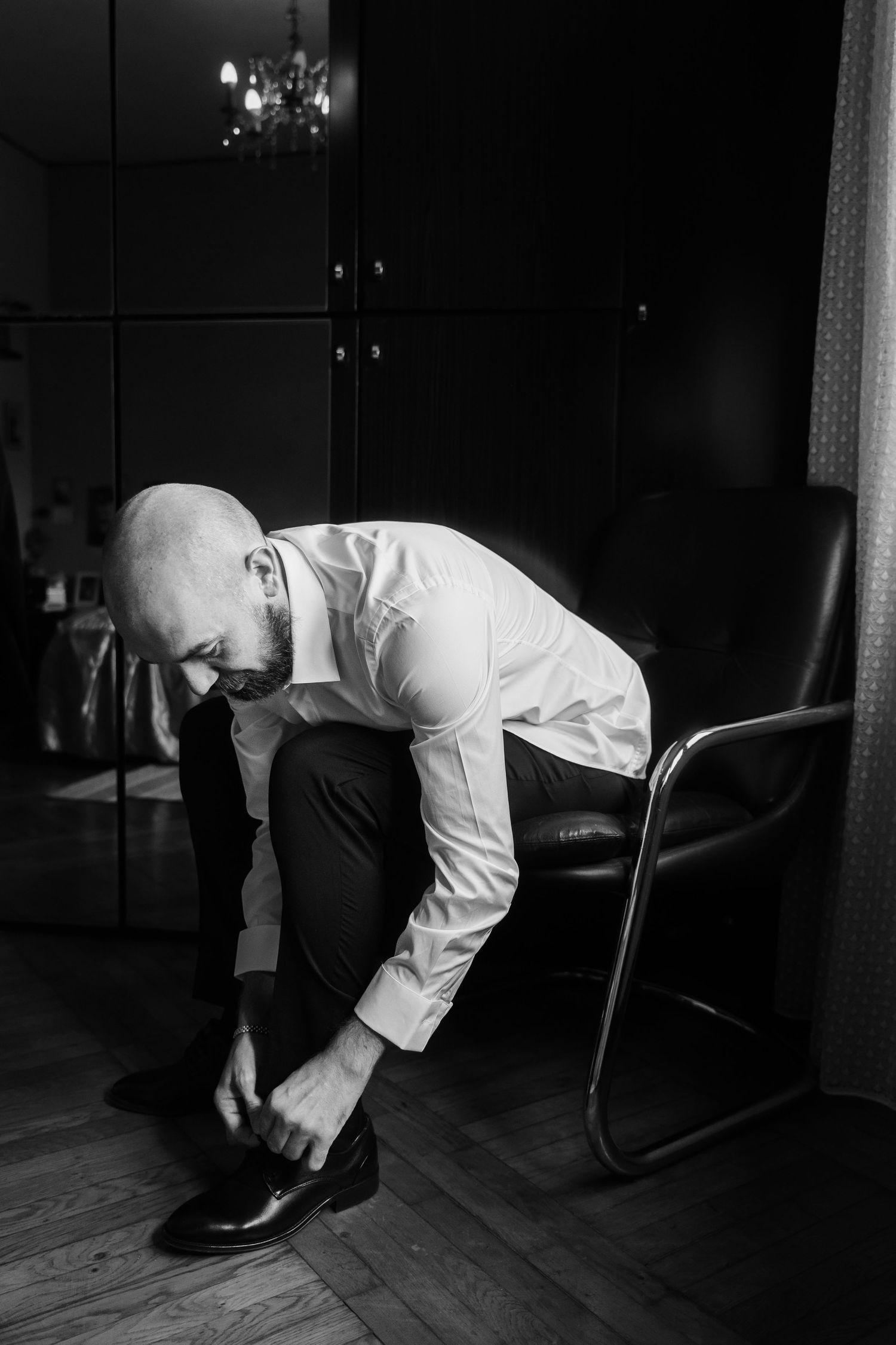 Bald man in white shirt and tie putting on dress shoes beside a leather chair in a dark room.