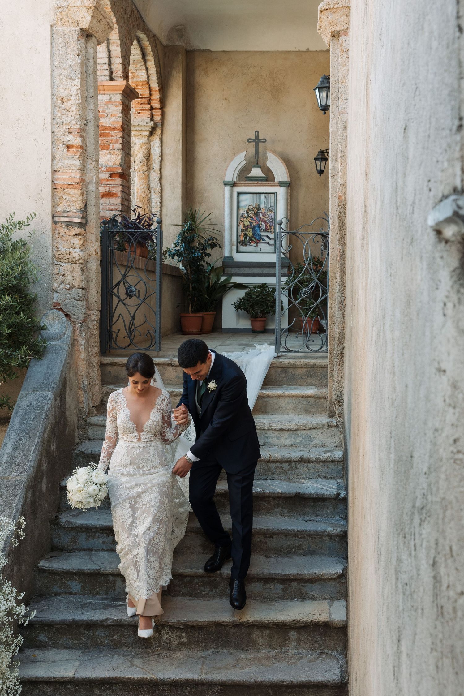 Bride and groom descending stone steps of historic Italian building holding bouquet.
