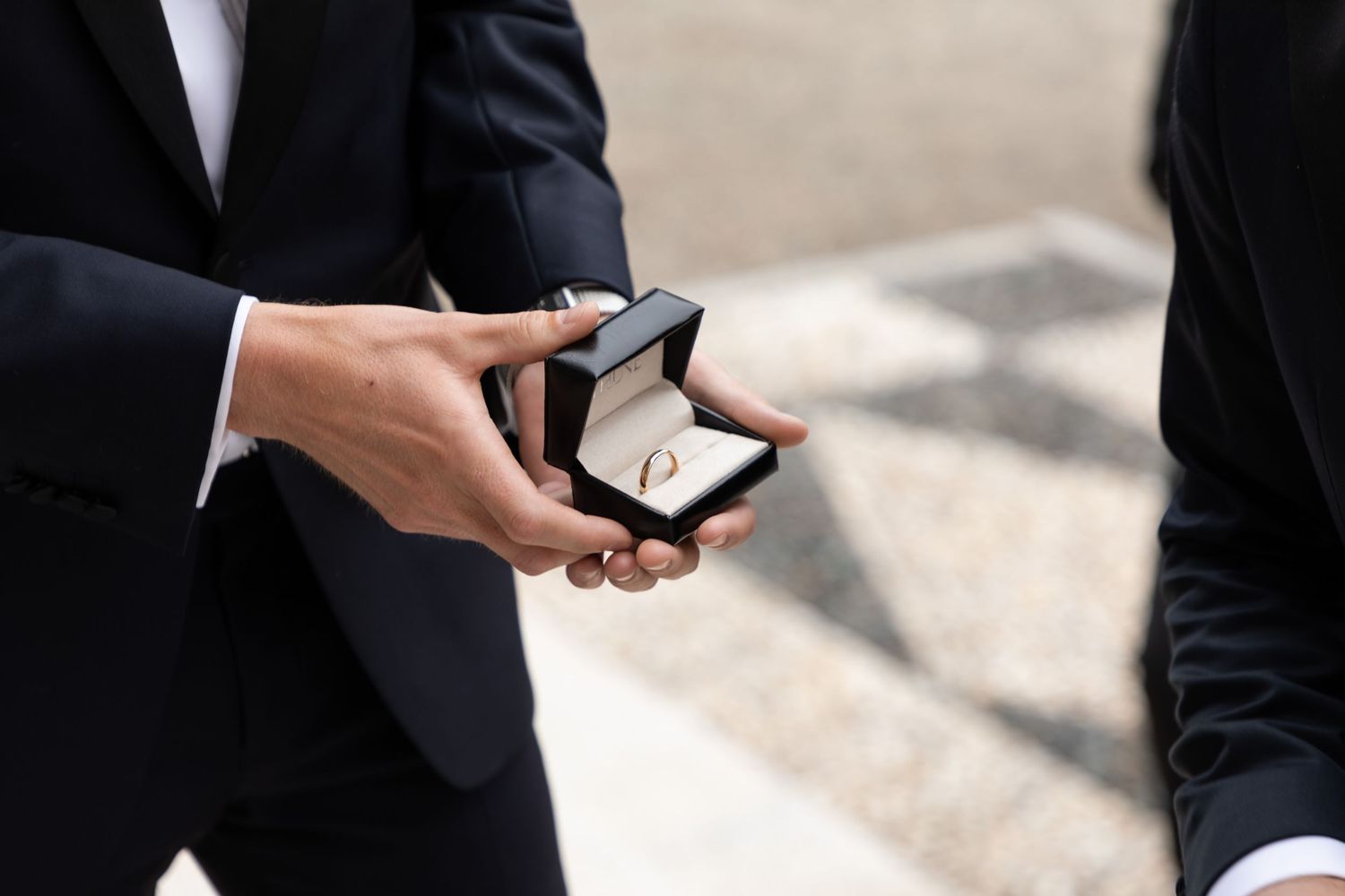 Close-up of two men in suits exchanging a wedding ring in an elegant black jewelry box outdoors.