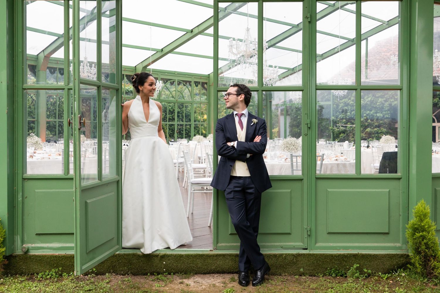 A bride and groom stand in a green greenhouse doorway, smiling at each other on their wedding day.