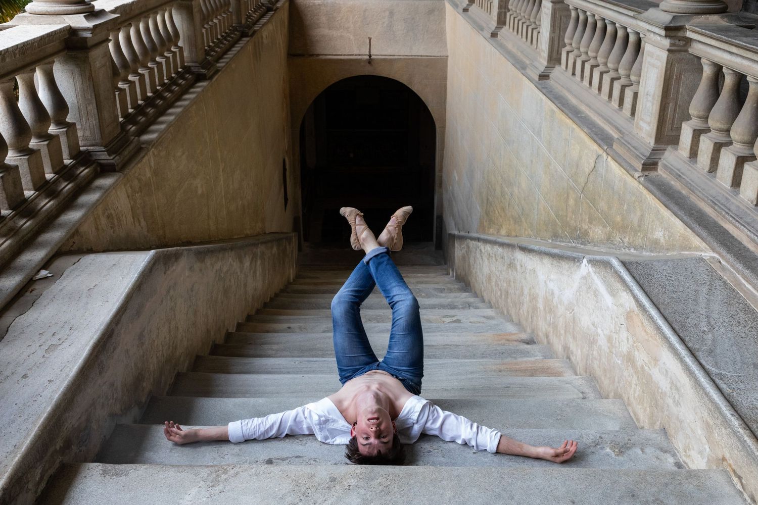 Ballet dancer lies dramatically spread-eagle on stone steps between ornate staircases of historic building.