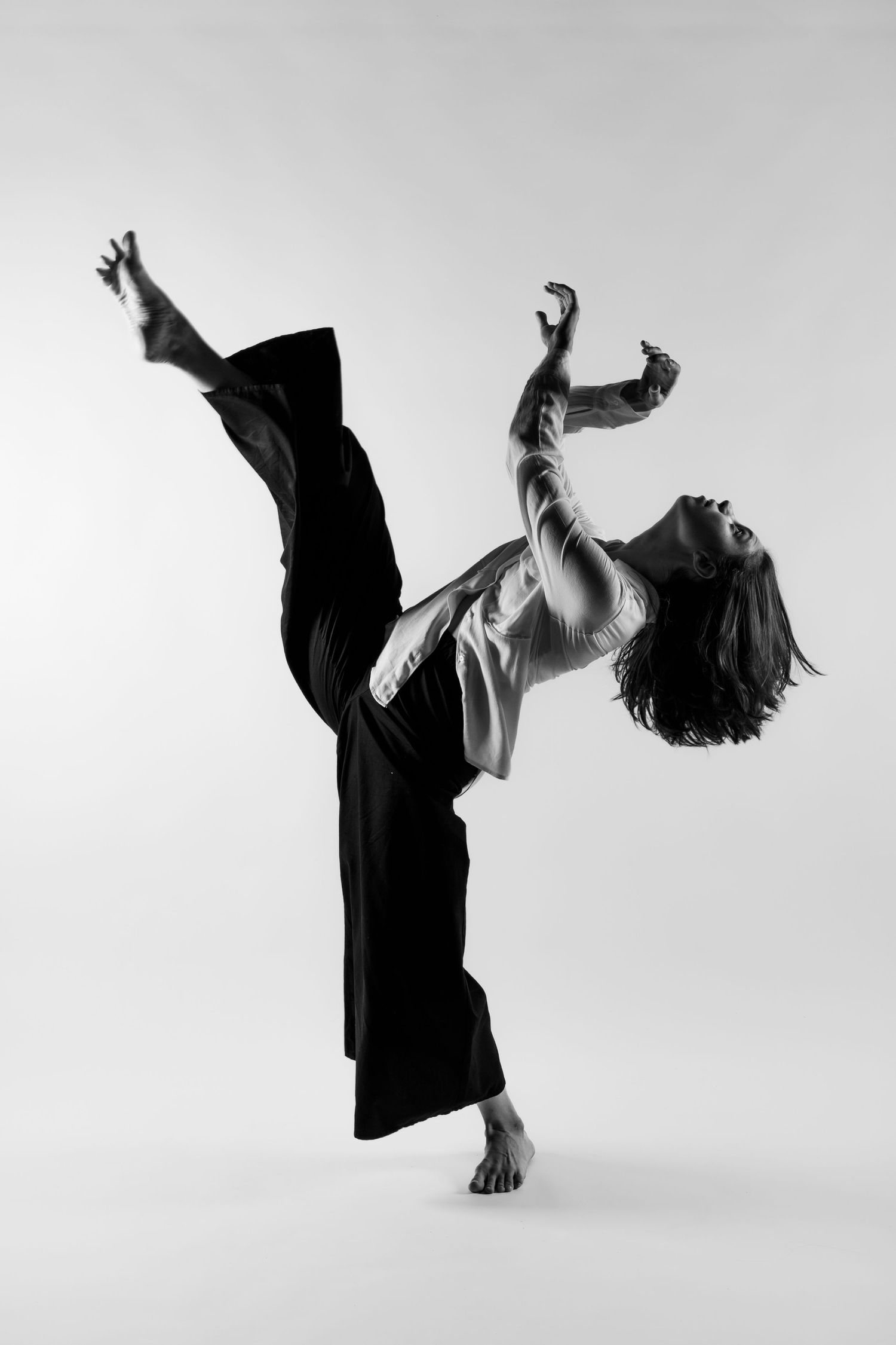 A barefoot dancer in black wide-leg pants executes a high kick with arms raised in a striking black and white studio shot.