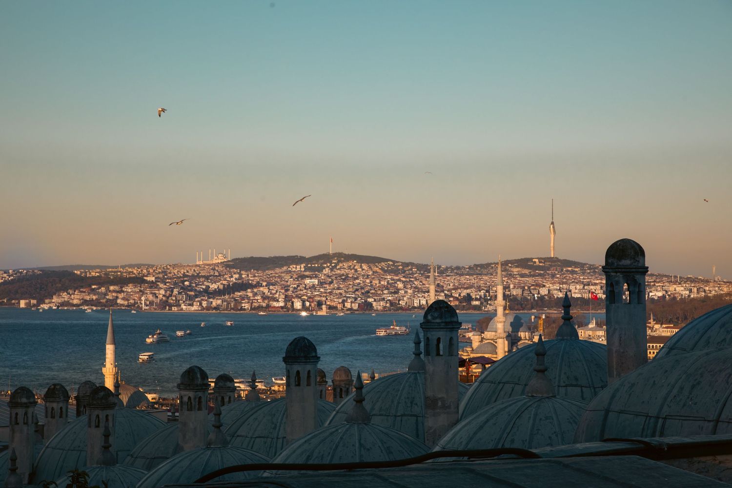 Panoramic sunset view of Istanbul skyline with mosque domes, minarets, and the Bosphorus strait in the background.