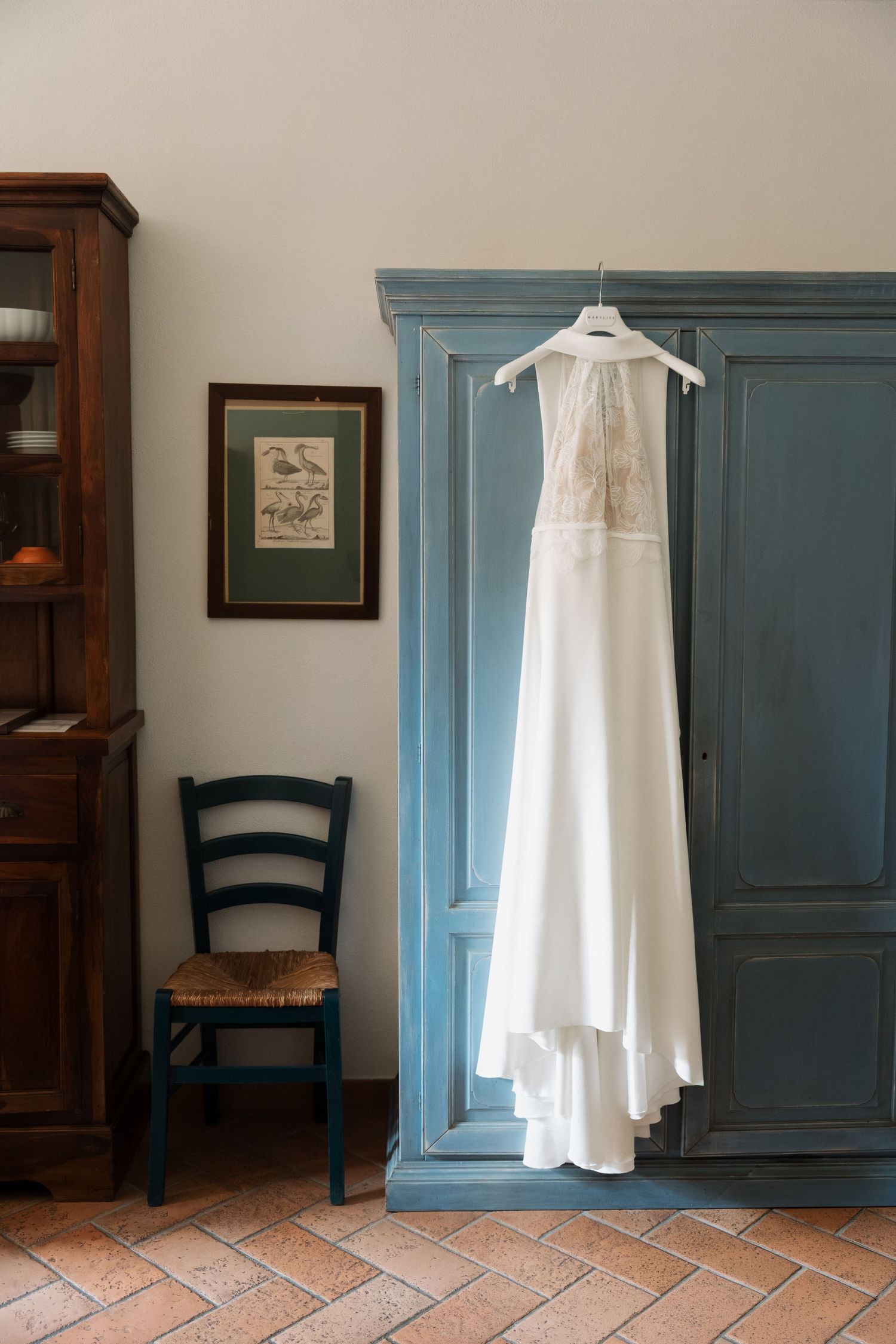 White wedding dress hanging on a blue armoire in a rustic room with terracotta floors and a wooden chair.