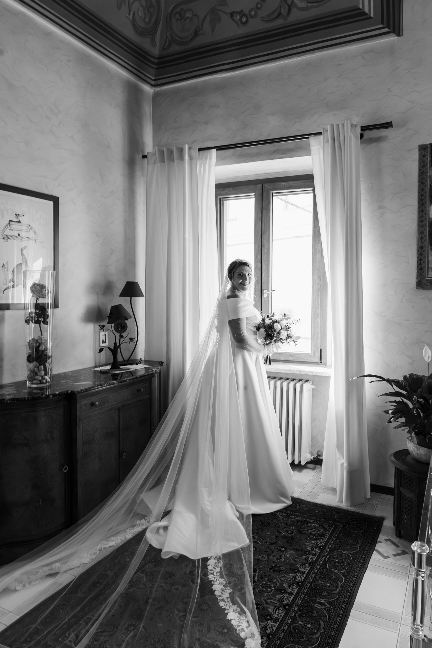 Black and white portrait of bride in strapless gown with long train standing by window.