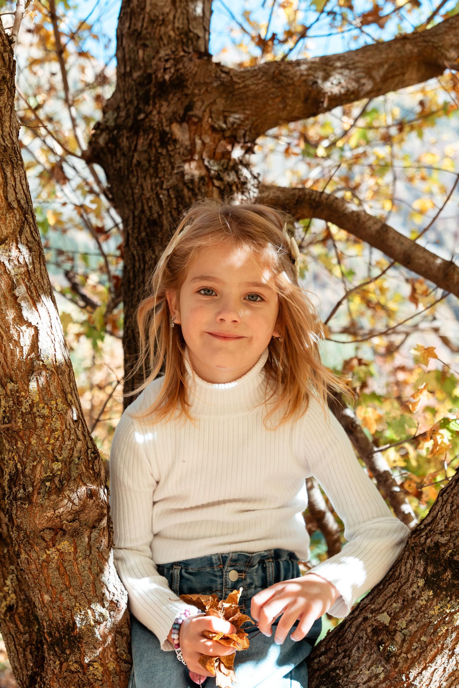 A young blonde girl in a white turtleneck sweater sits in an autumn tree holding a dried leaf.