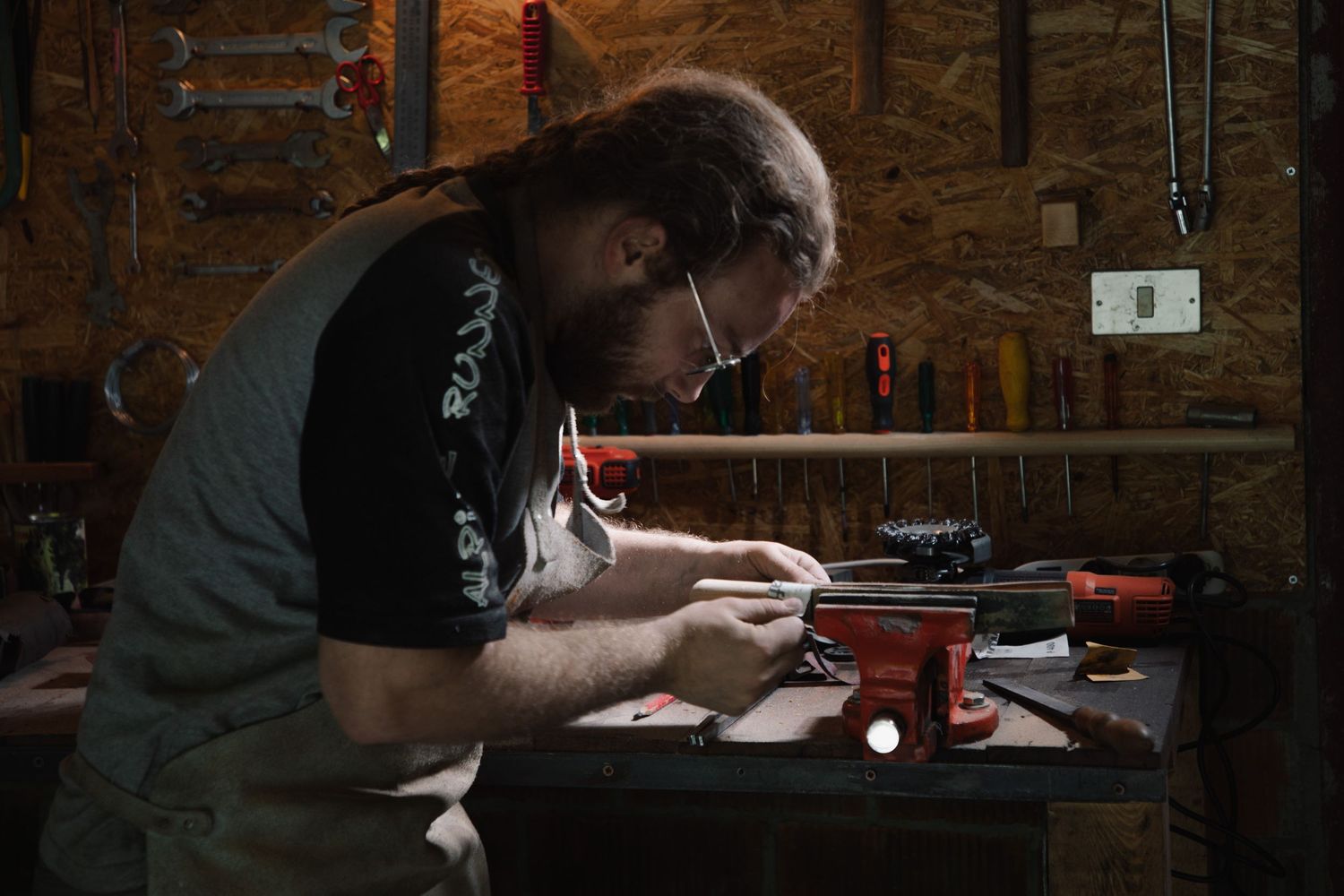 Long-haired craftsman in leather apron carefully crafting a knife at a dimly lit workshop bench.