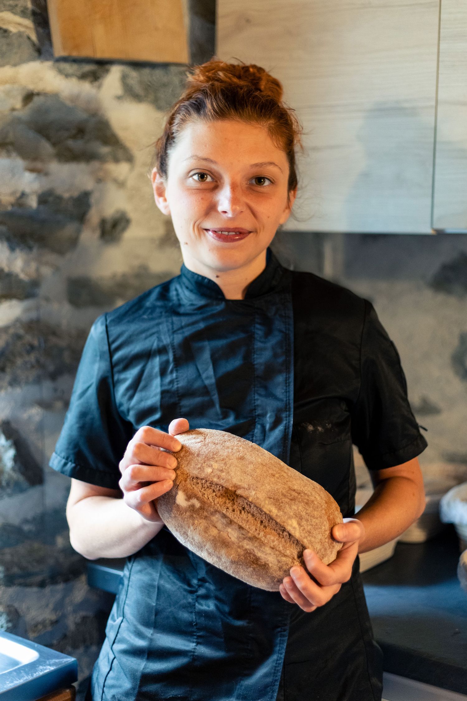 Female chef in black uniform proudly holding freshly baked artisan sourdough bread loaf.