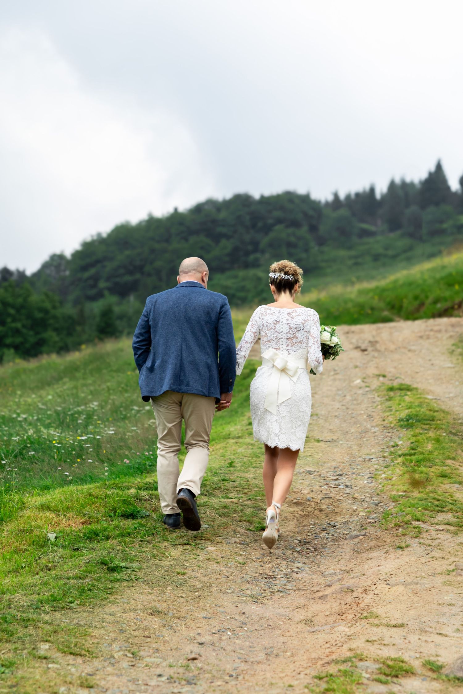 Bride and groom walk hand in hand along a rural dirt path through green meadows and forest hills.