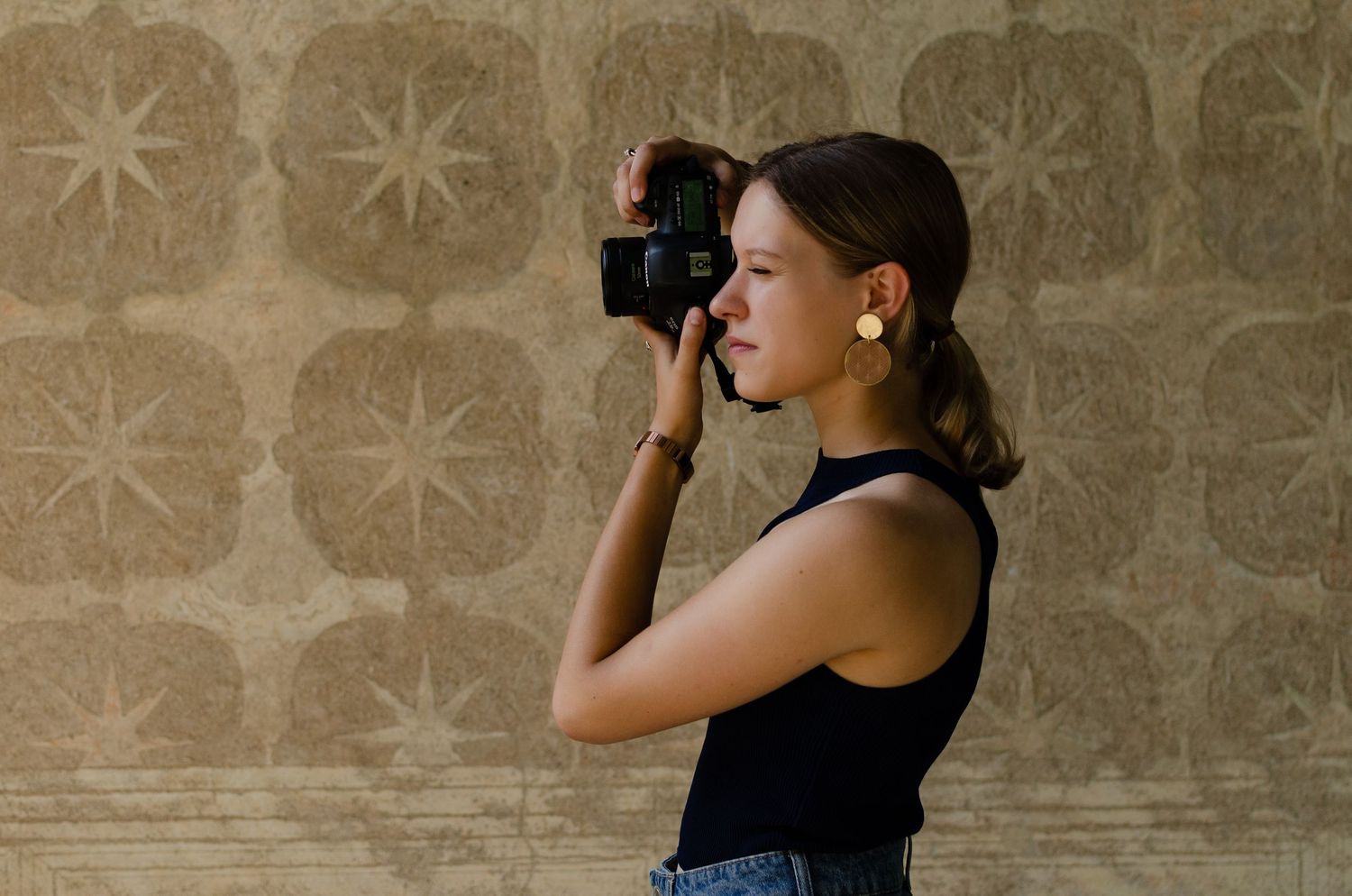 Young woman photographer capturing photos with DSLR camera against decorative patterned wall background.