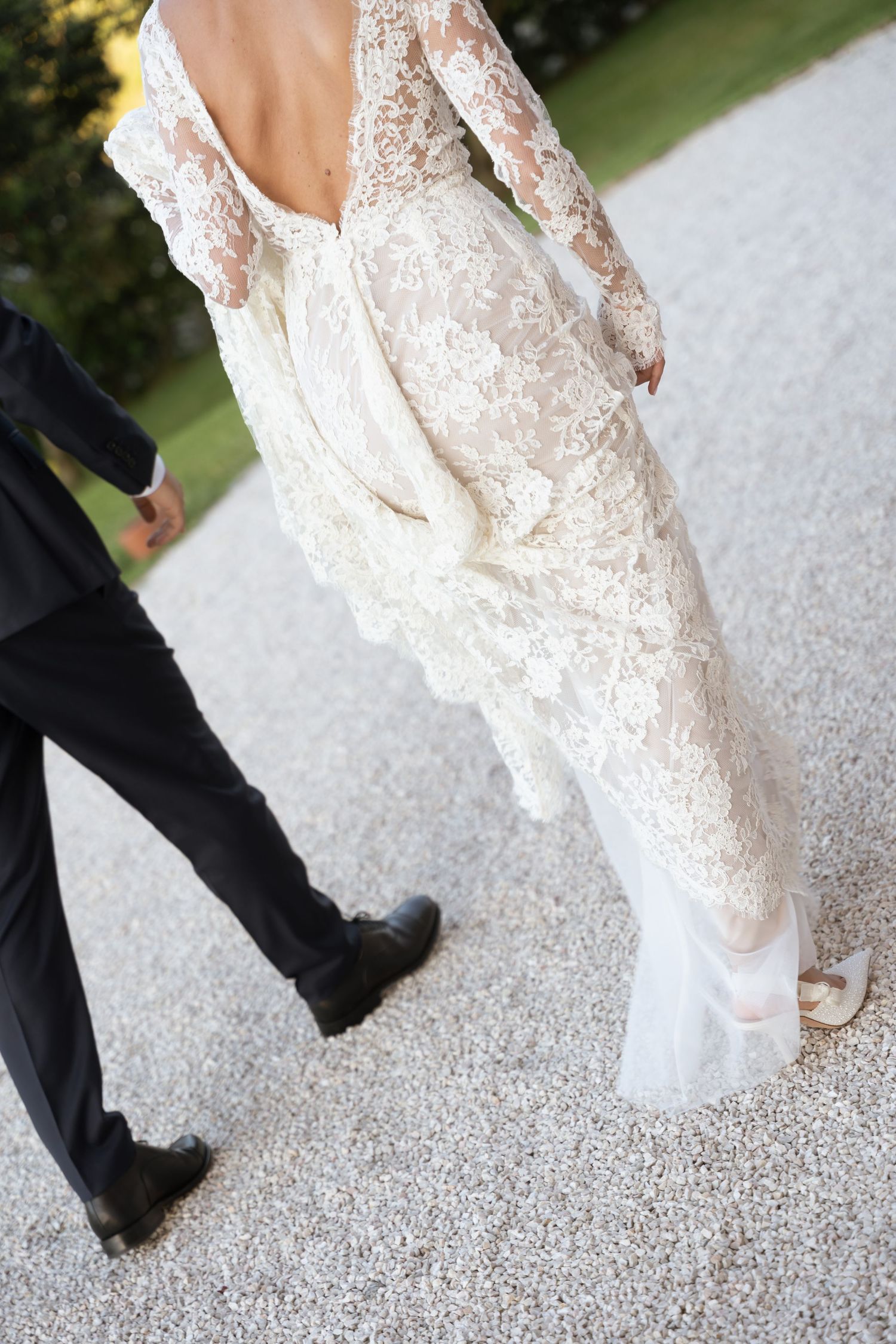 Bride in elegant long-sleeve lace wedding gown with open back walks beside groom in black suit on gravel path.