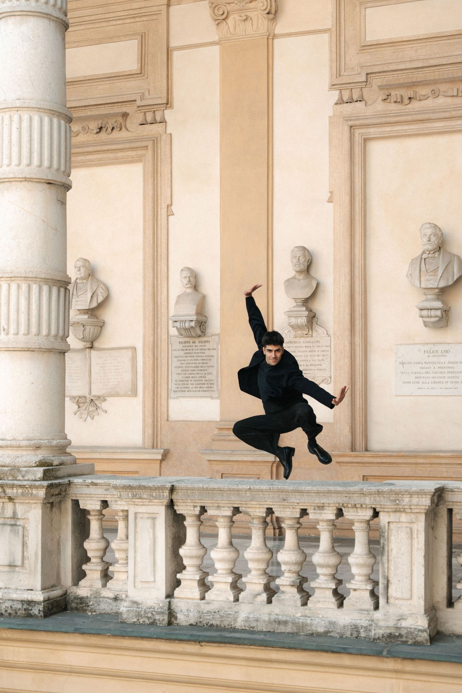 Male dancer in black leaping over white balustrade in classical building with marble busts.