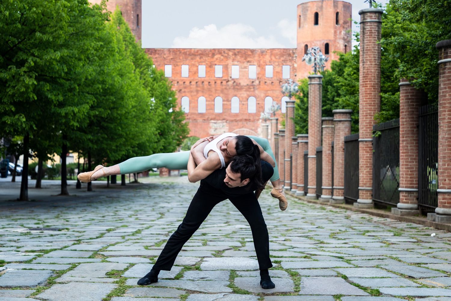 Male dancer performs dramatic acrobatic lift with female dancer outdoors near ancient Roman ruins.
