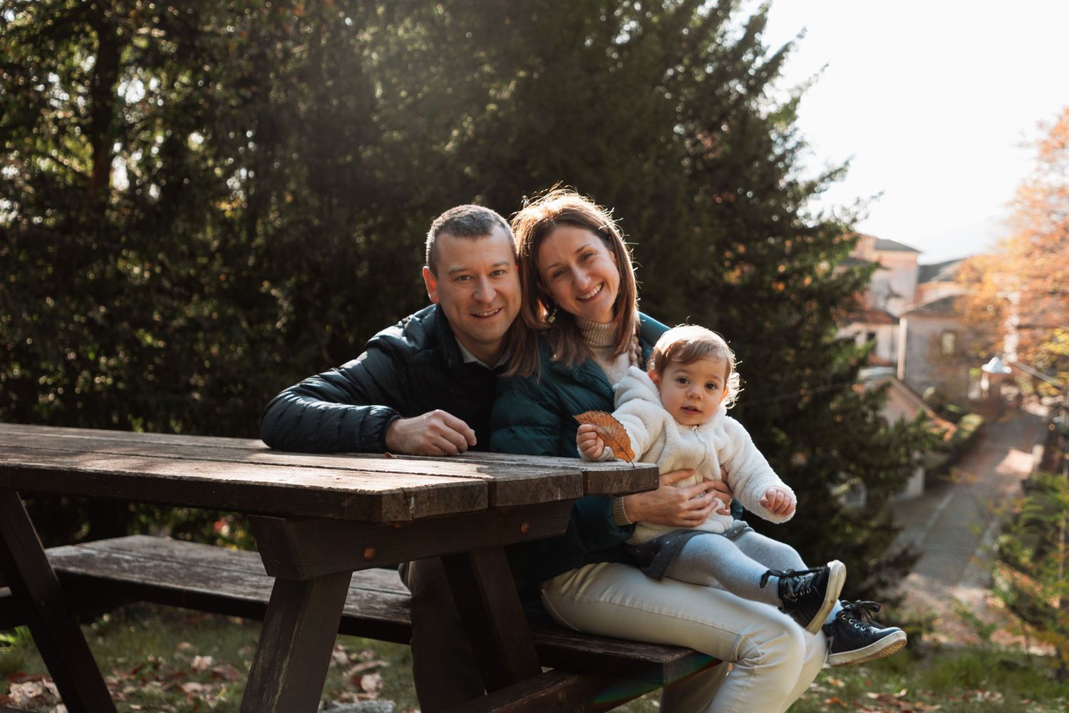 A happy couple sits at an outdoor wooden table with their baby in a sunny garden setting.