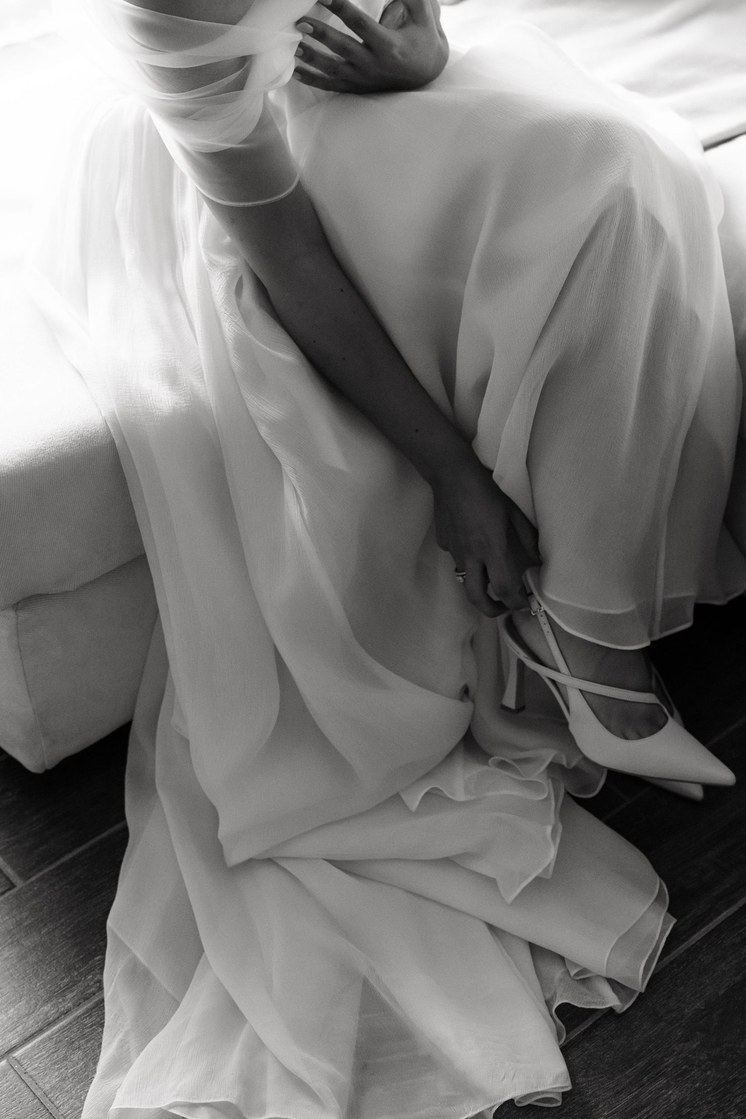 Black and white photo of a bride in a flowing white gown holding heels while sitting on the floor.