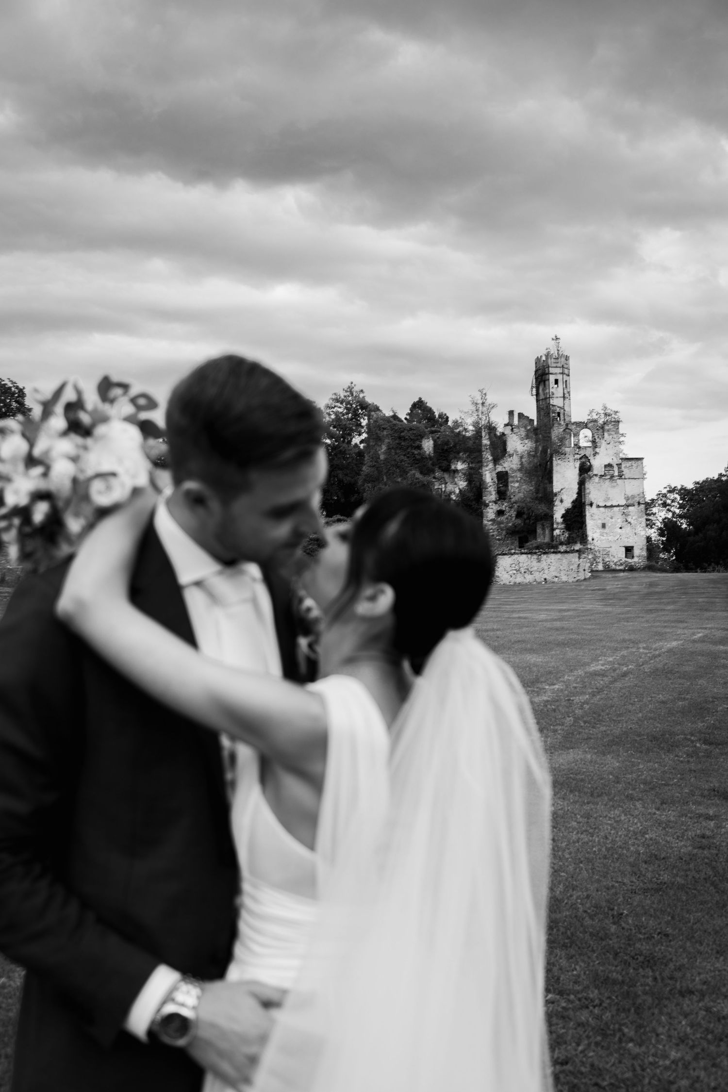 Black and white photo of a bride and groom sharing a kiss with a historic castle ruin in the background.