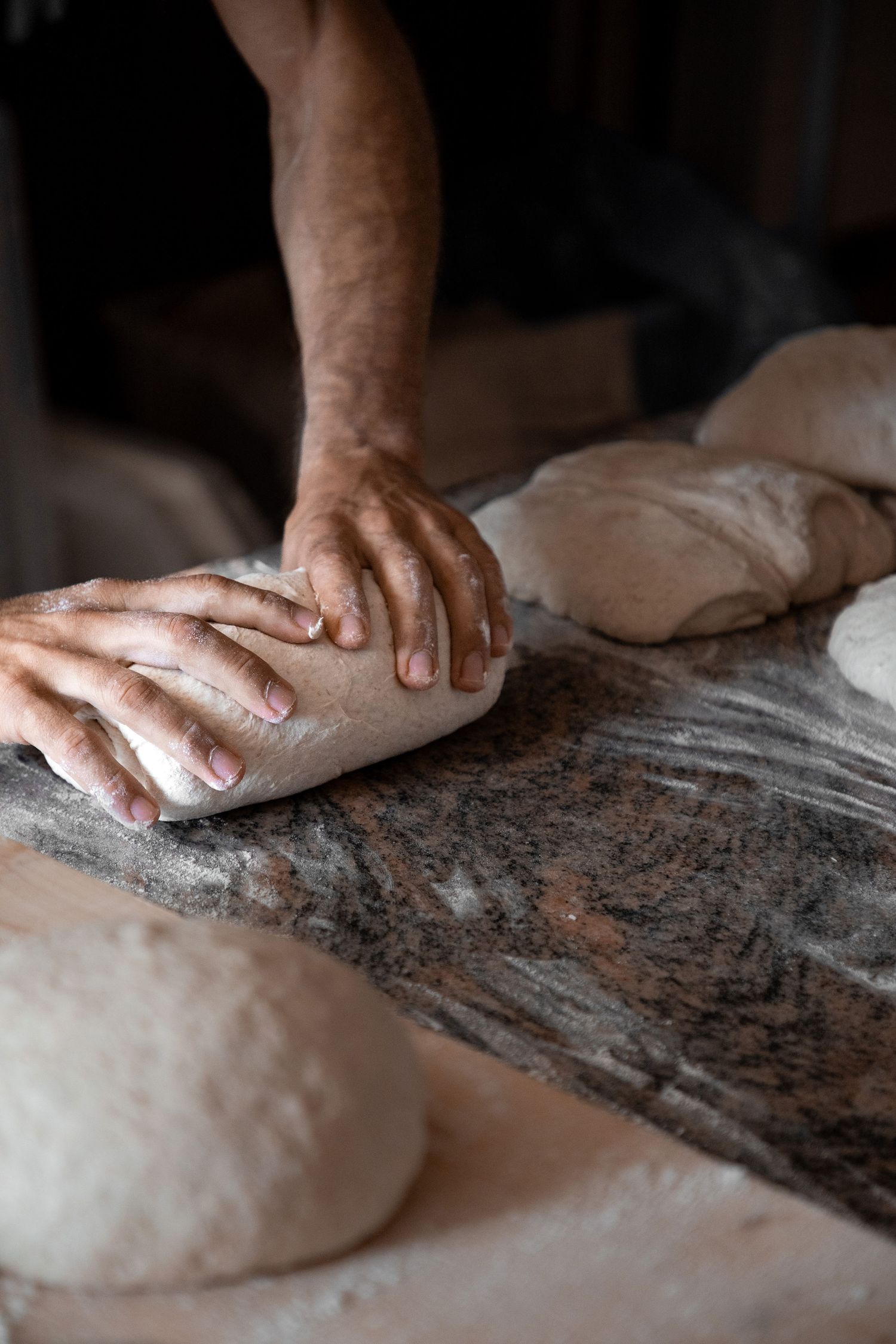 A baker kneading bread dough on a flour-dusted marble countertop with multiple dough portions nearby.