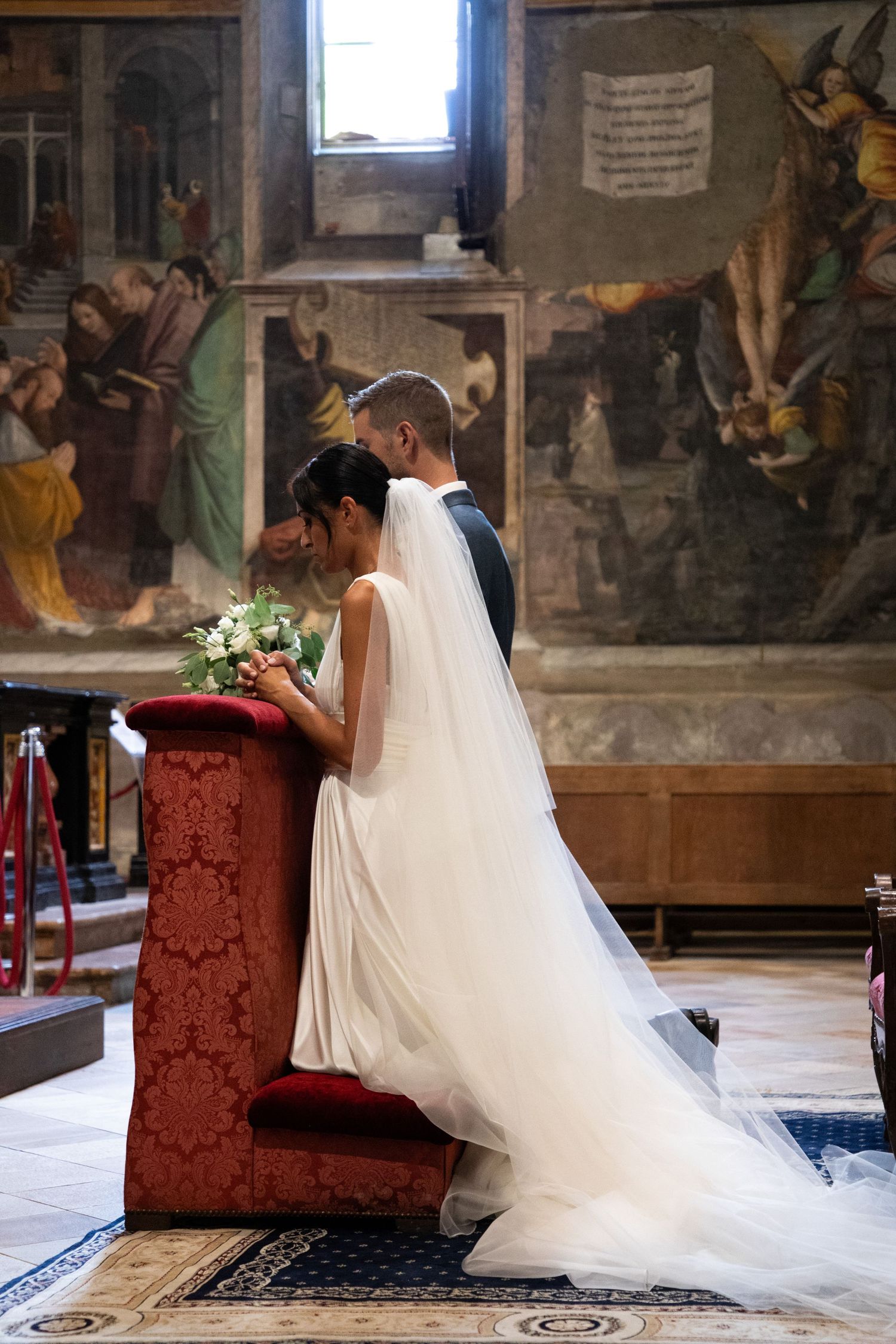 Bride in flowing white gown and veil stands with groom at ornate red kneeler in frescoed church.