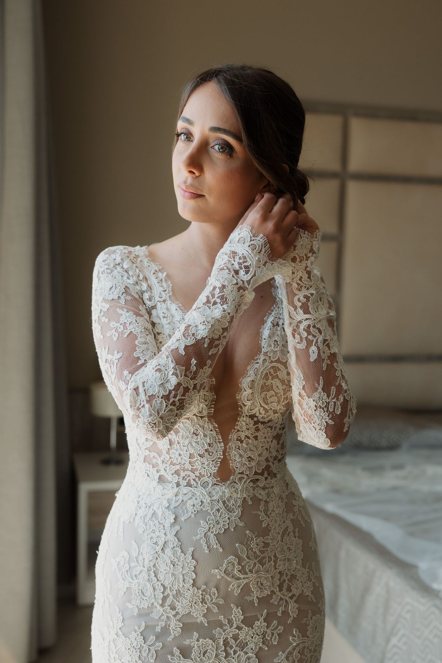 Bride in elegant long-sleeve lace wedding gown adjusting earring in softly lit bedroom.