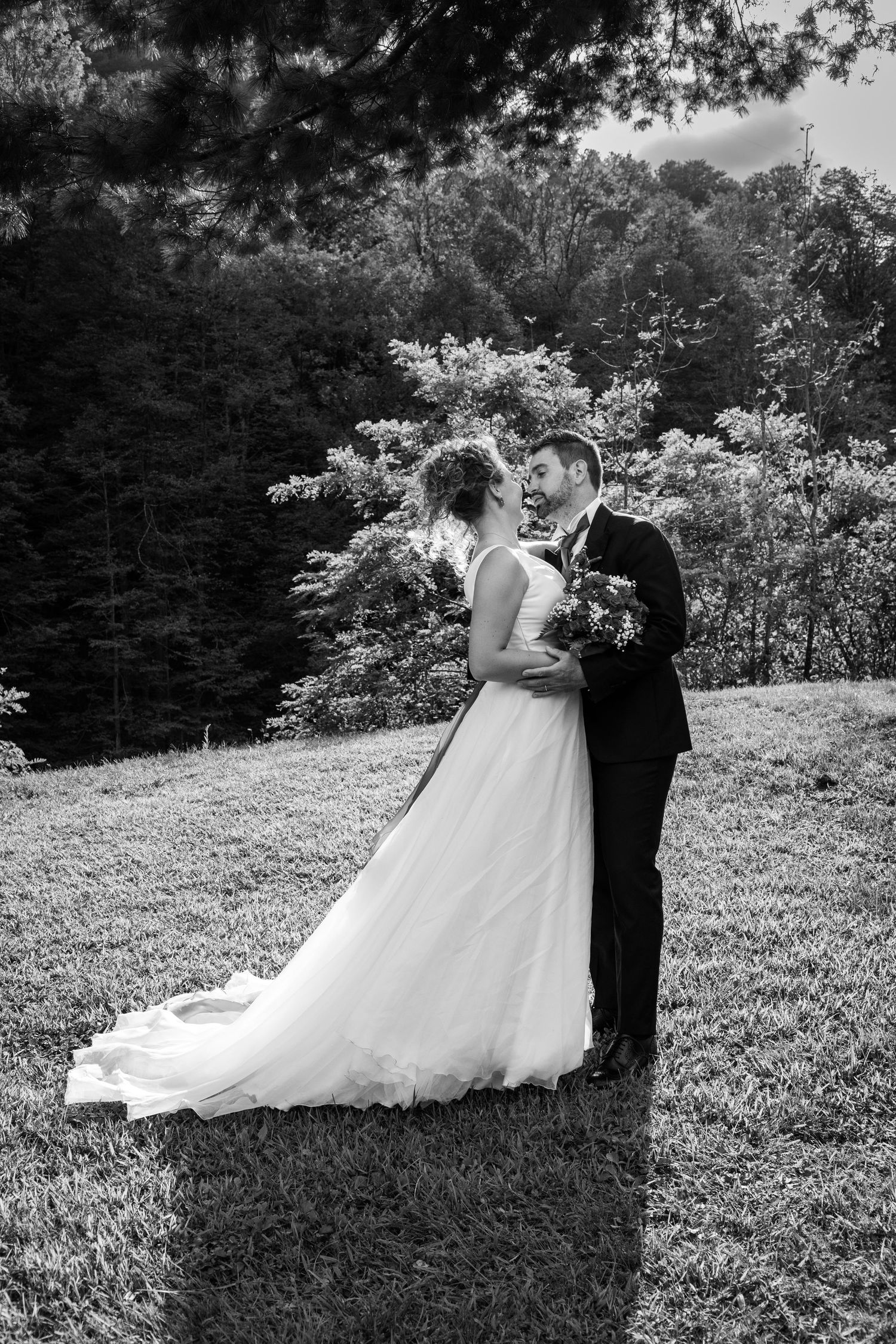 Black and white portrait of bride and groom embracing on hillside surrounded by lush forest.