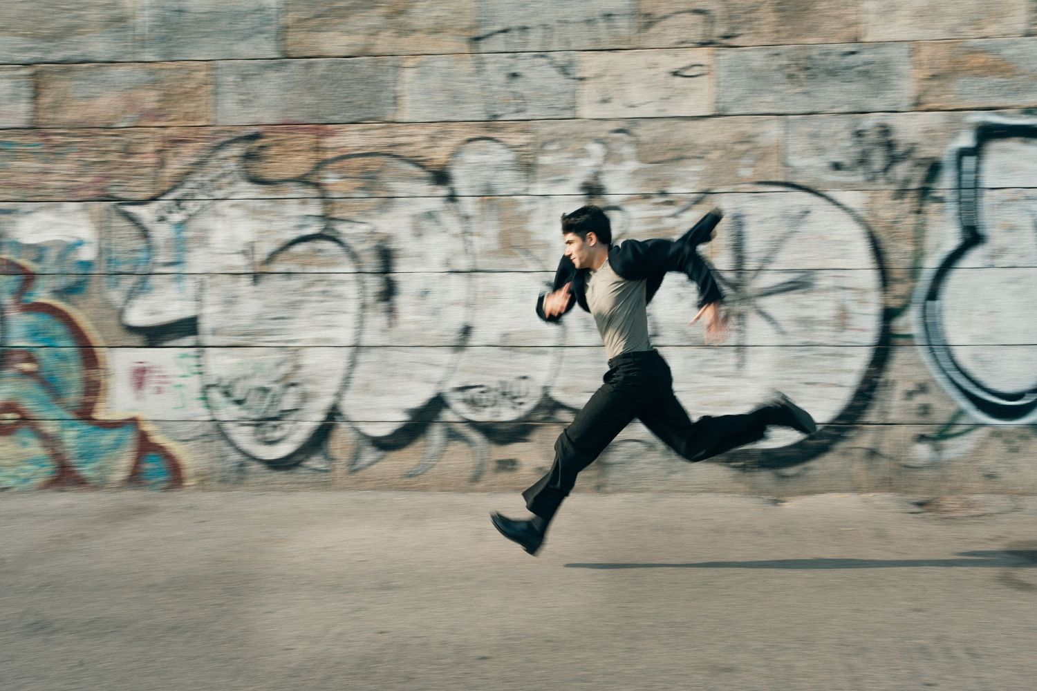 Male dancer in black running dynamically past colorful graffiti-covered urban wall.