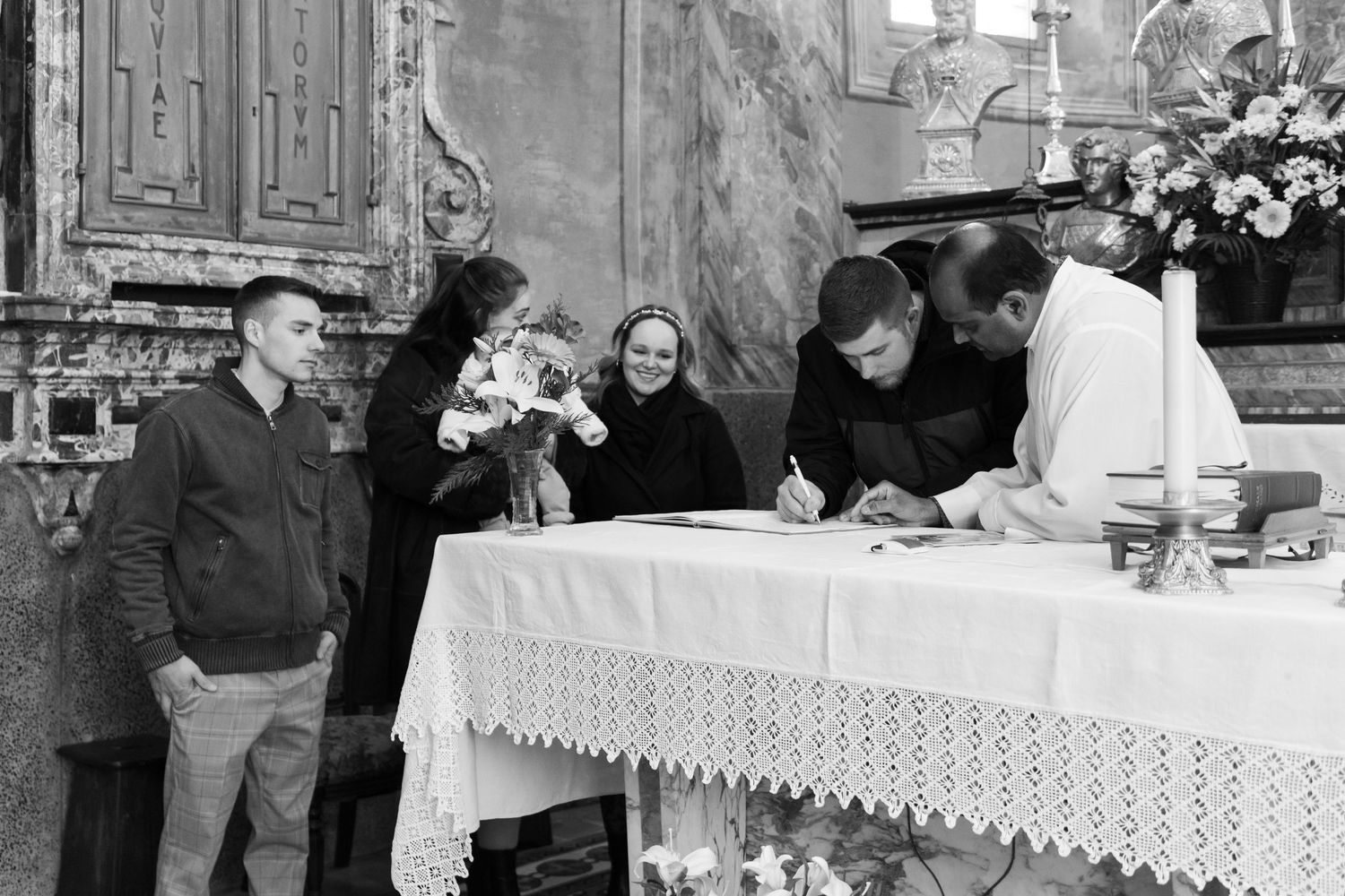 Black and white photo of family signing baptism documents at a church altar with floral decorations.