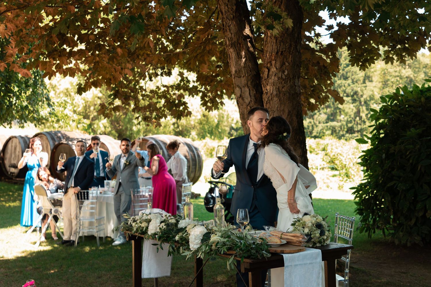 Bride and groom share a tender moment at outdoor wedding reception under autumn tree with guests in background.