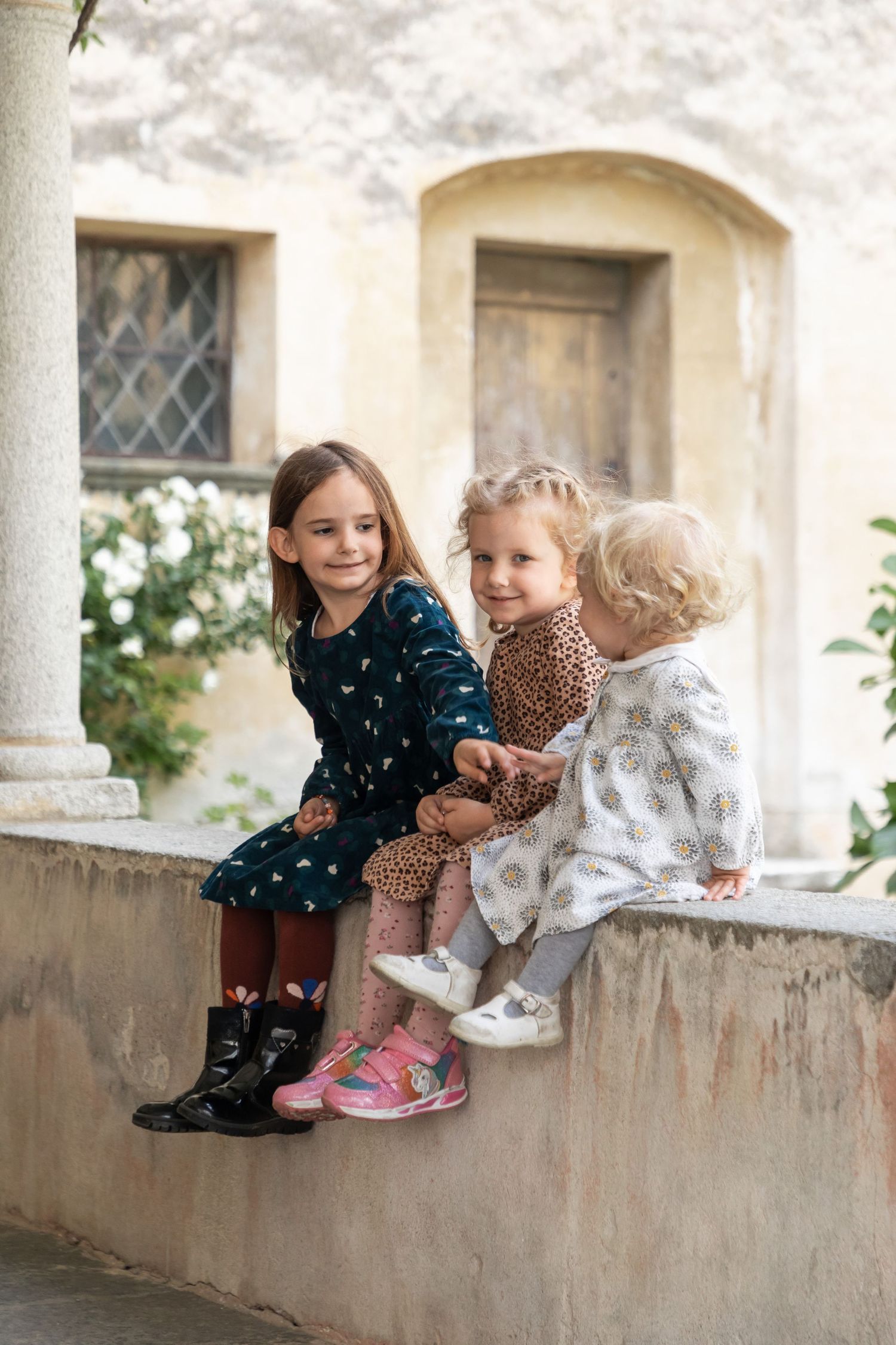 Three young girls in colorful dresses sitting on stone wall outside rustic European building.