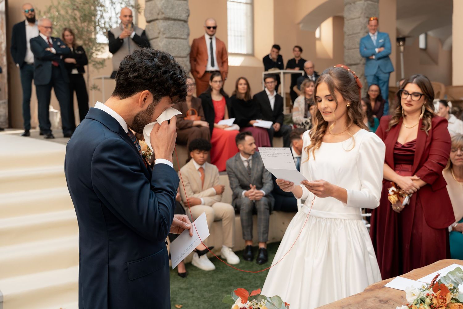 Emotional groom wiping tears as bride reads vows during outdoor wedding ceremony.
