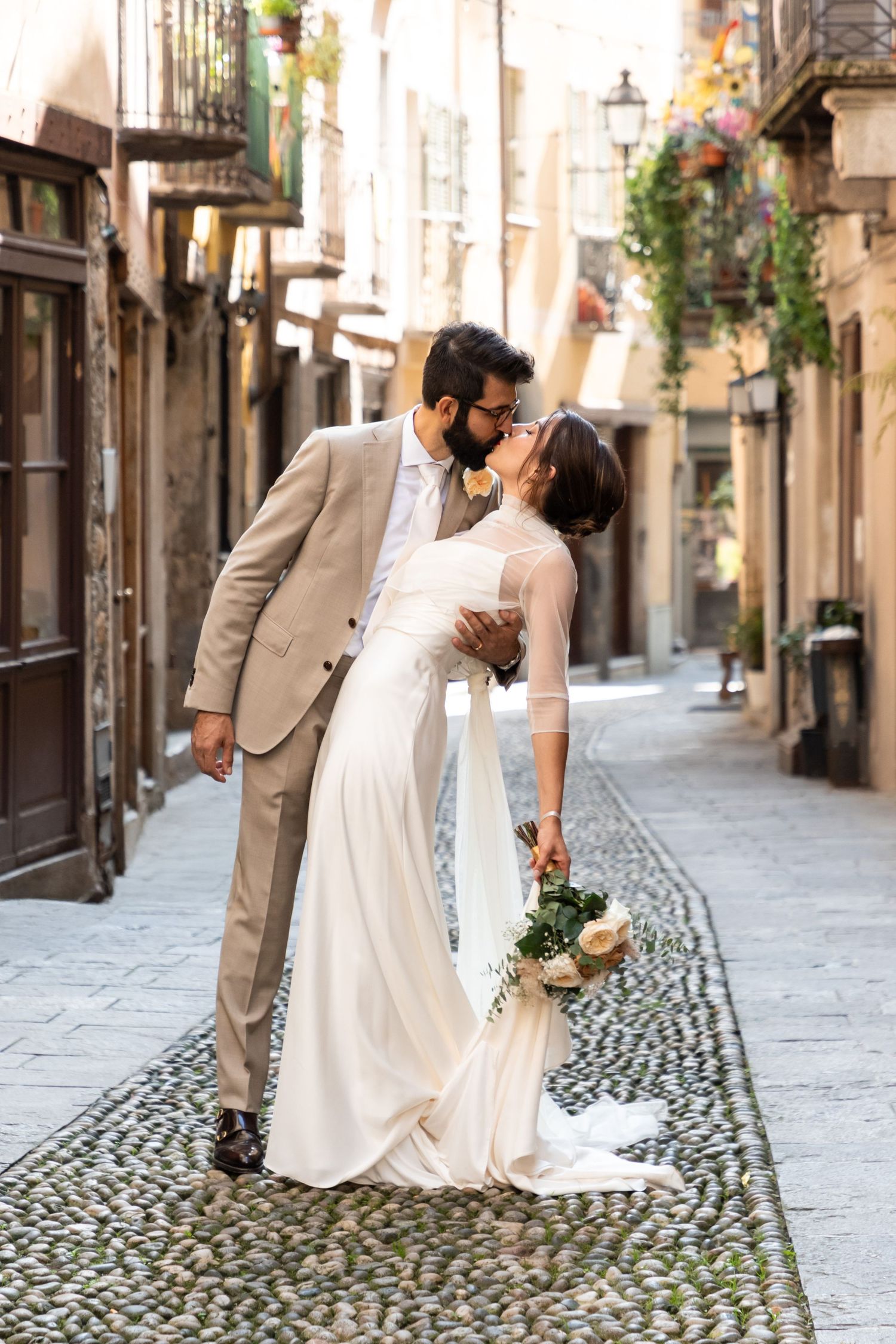 Groom dips bride for a kiss on charming cobblestone street lined with old European buildings.
