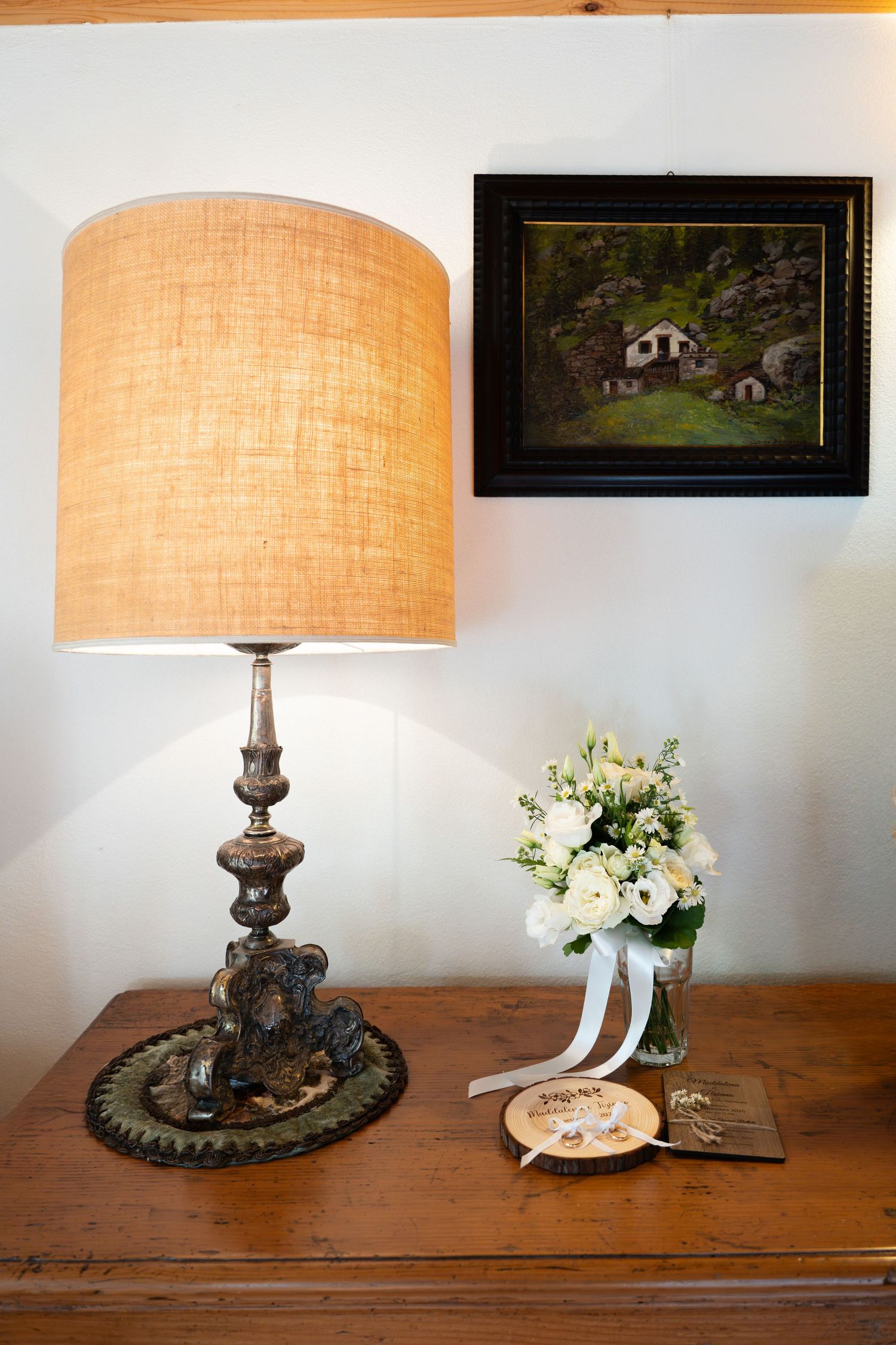 Vintage lamp beside white rose bridal bouquet and jewelry on rustic wooden dresser.