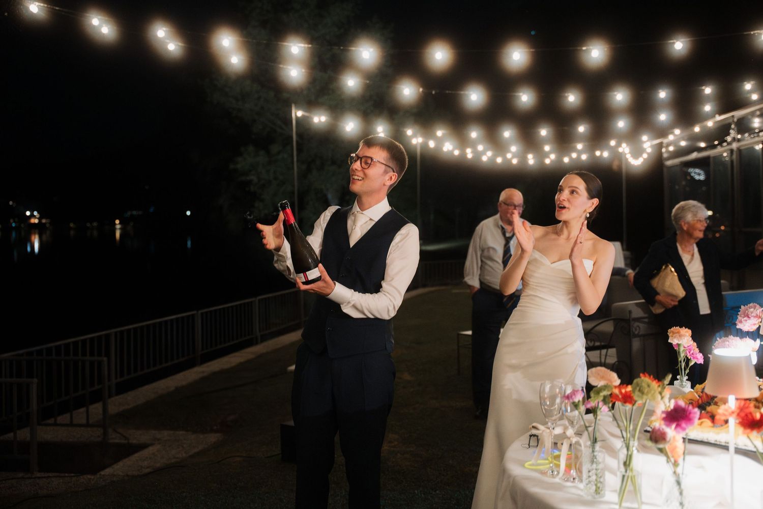 Bride and groom toast with champagne beside a decorated wedding cake at an outdoor evening reception.