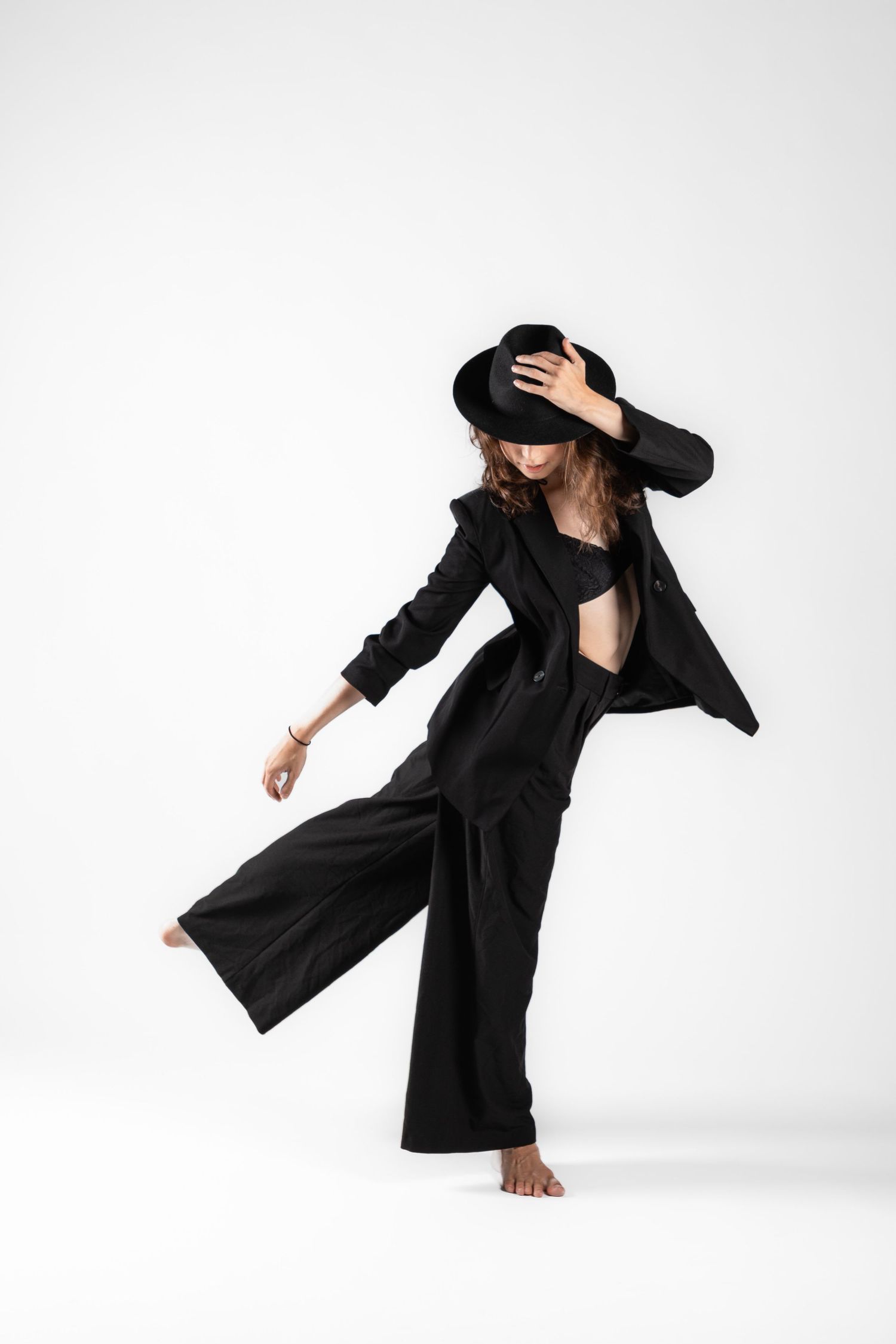 A woman in a black suit and wide-brim hat poses dynamically, lifting one leg while touching her hat against a white background.