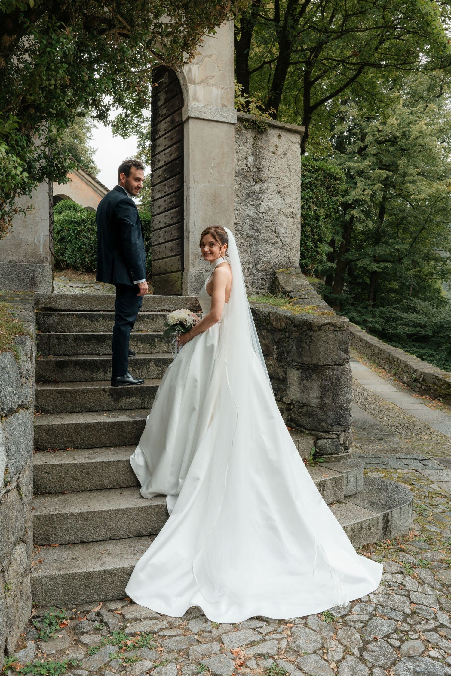 Bride in flowing white gown with veil looks back on stone steps as groom stands in background.