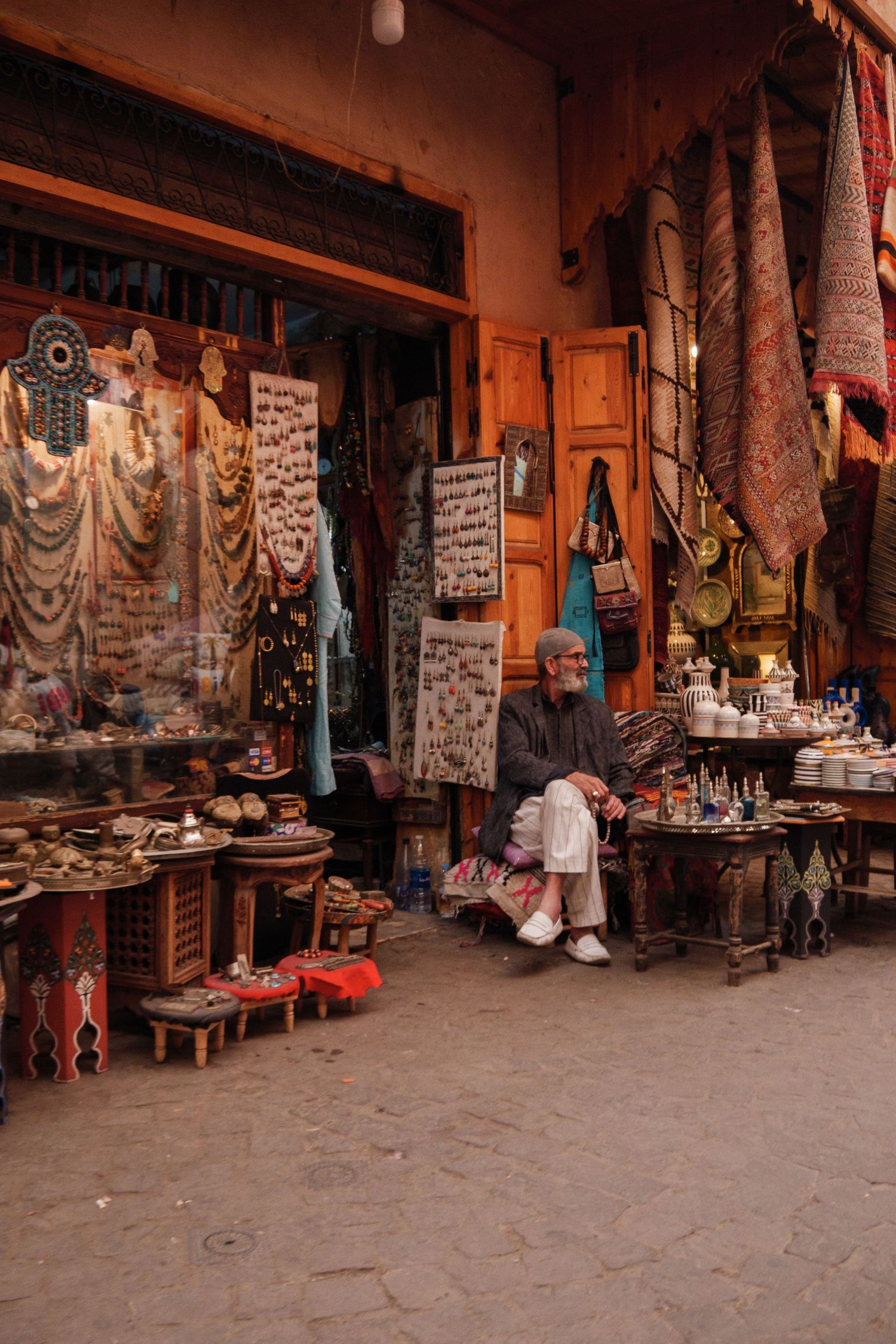 Elderly vendor selling traditional Moroccan crafts, jewelry, and rugs in a colorful medina market.