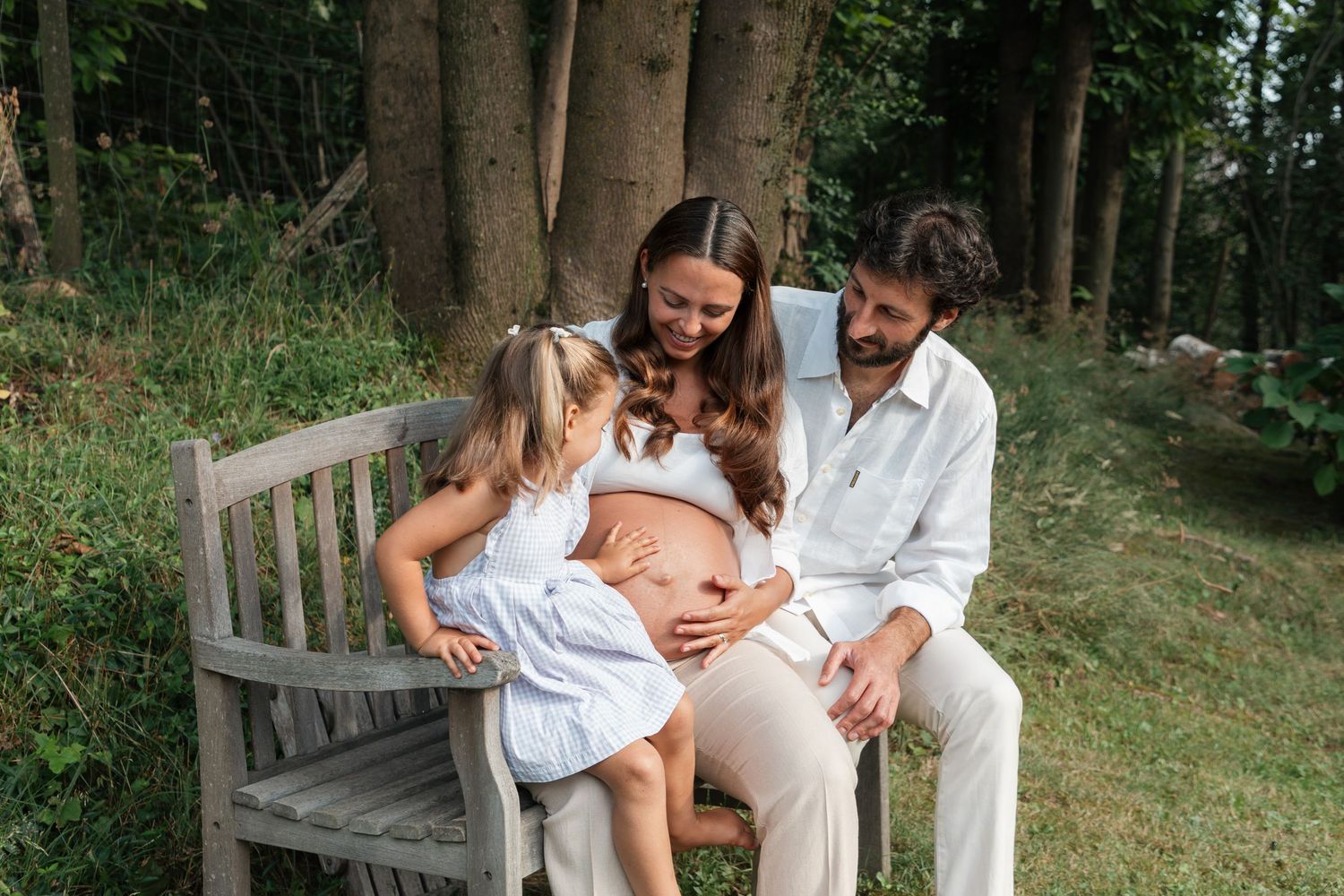 A happy family with a pregnant mother, father, and young daughter sitting on a wooden bench in a lush garden.