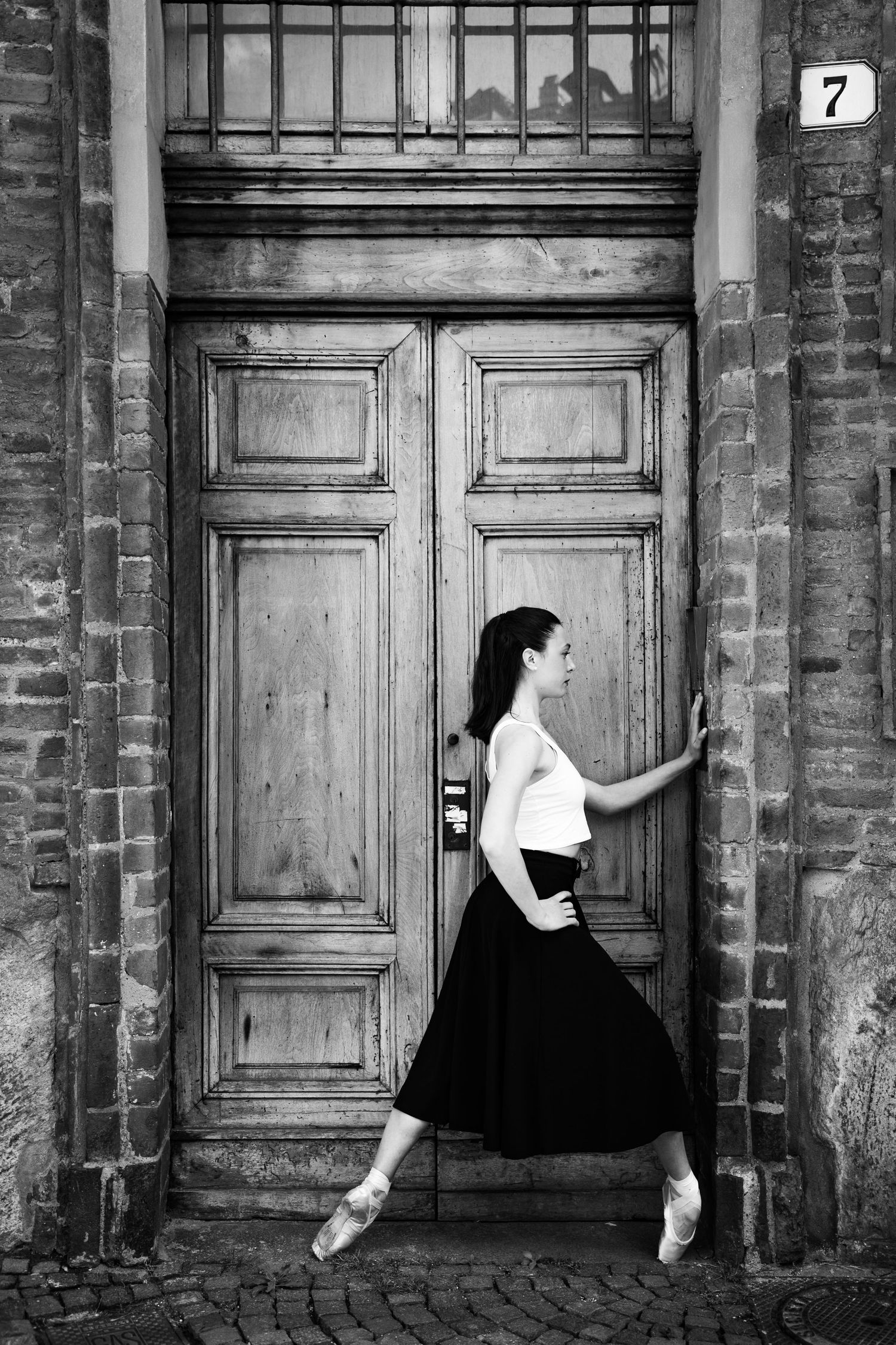 Ballerina in black skirt and pointe shoes posing by vintage wooden door number 7, black and white.