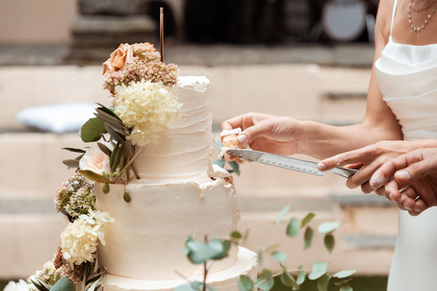 Bride cutting beautiful three-tier white wedding cake decorated with fresh flowers and greenery.