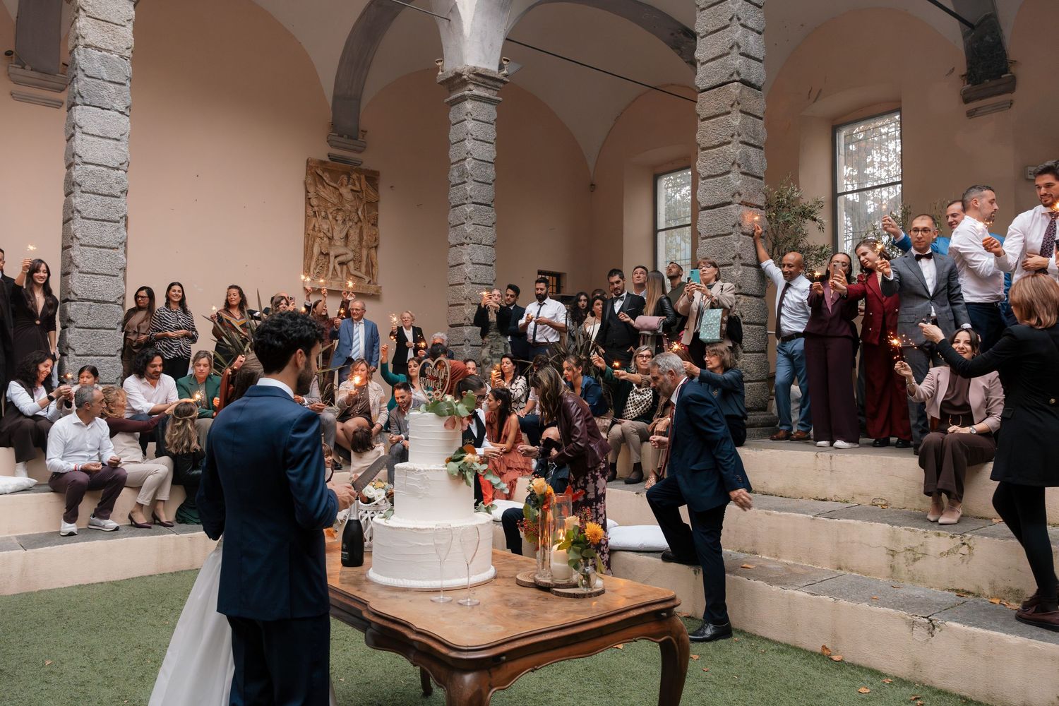 Wedding guests gather around tiered cake table in stunning arched stone venue during cake cutting celebration.