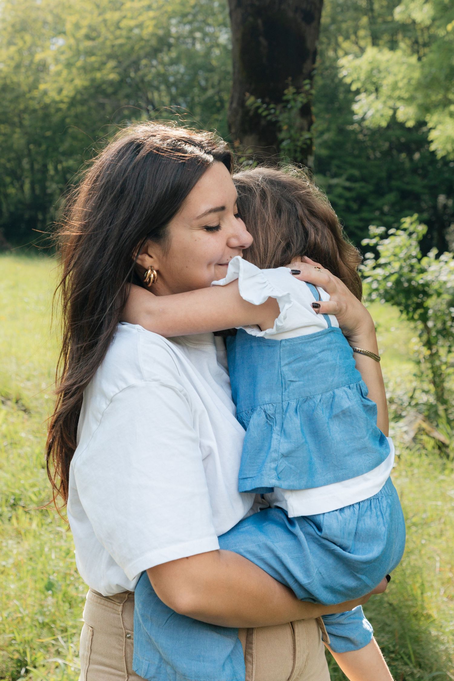 A dark-haired mother tenderly embraces her young daughter in a blue dress outdoors.