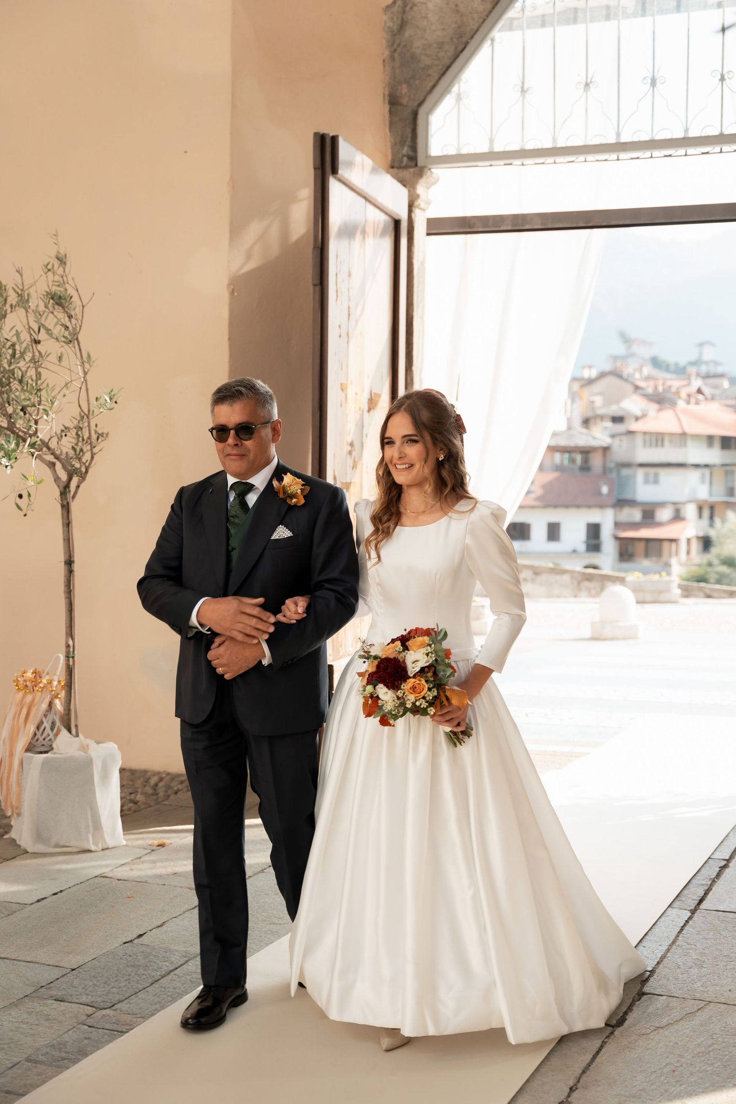 Smiling bride in long-sleeve white satin gown walks down aisle on father's arm holding colorful bouquet.